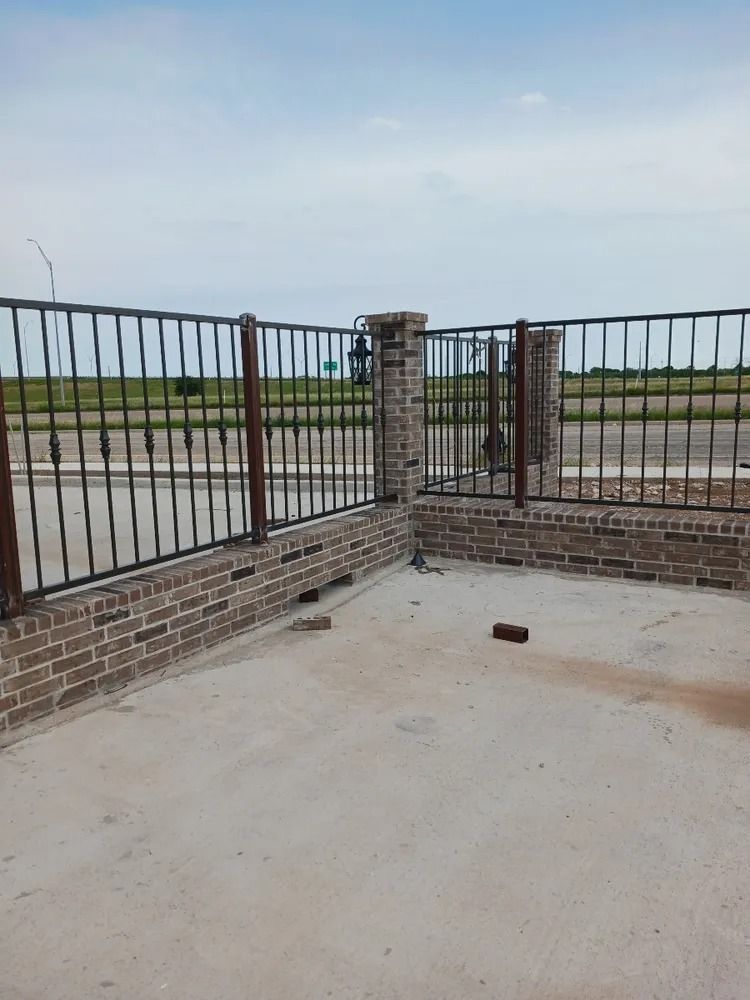 Brick and wrought iron fence with concrete ground against a field and sky background.