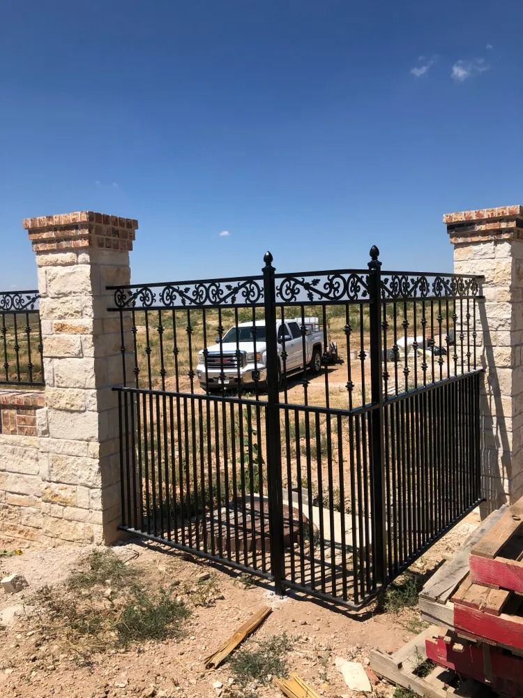 Black wrought-iron gate with stone pillars; white truck in the background, blue sky.