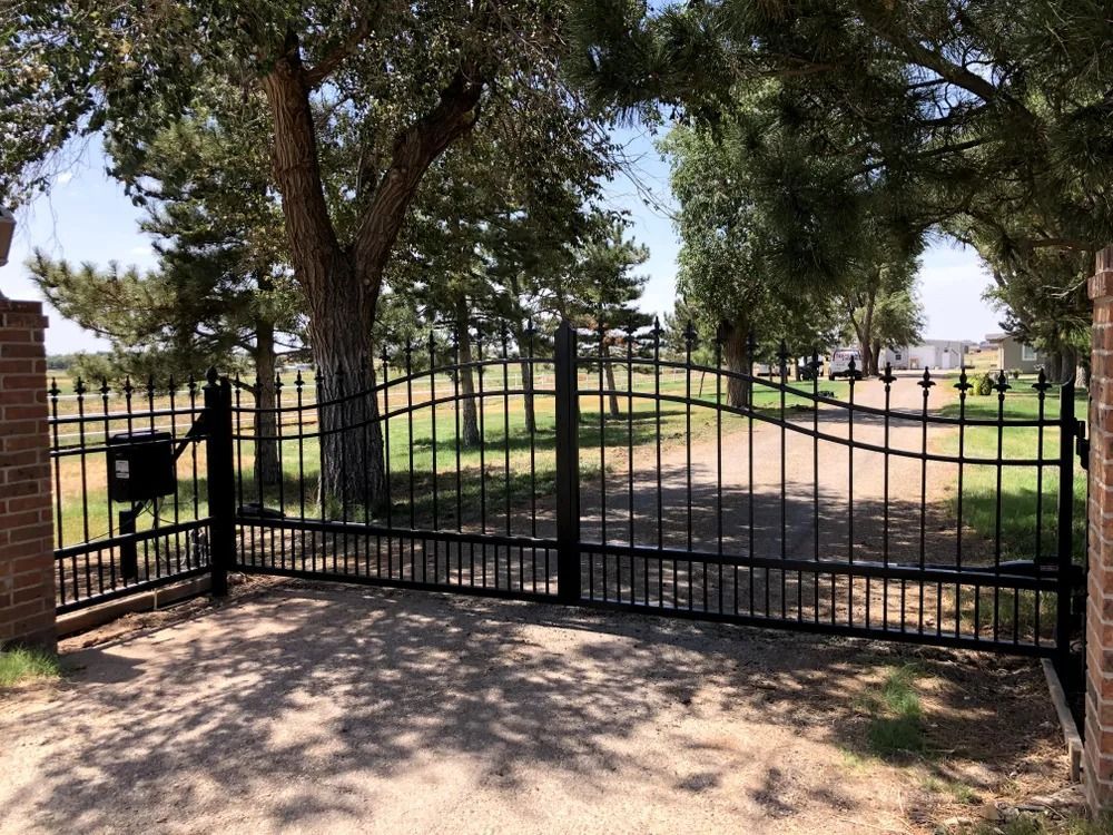 Black metal driveway gate in front of a house. Tall trees and brick pillars flank the gate.