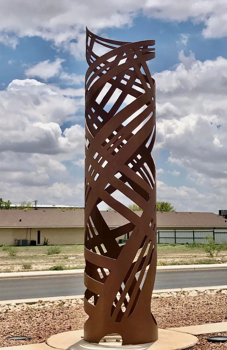 A tall, brown metal sculpture with interwoven ribbons stands outdoors in front of a building, under a cloudy sky.