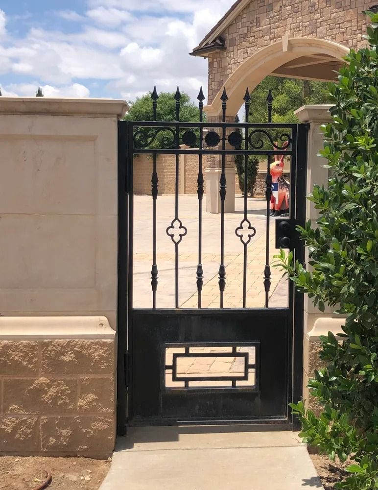 Black wrought-iron gate set in a tan stone wall, with a concrete walkway leading up to it, next to green bushes.