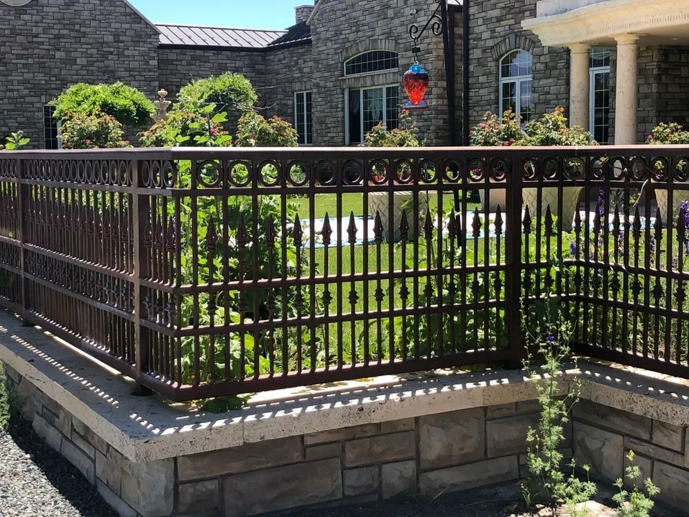 Brown metal fence atop a stone wall, in front of a house and garden.