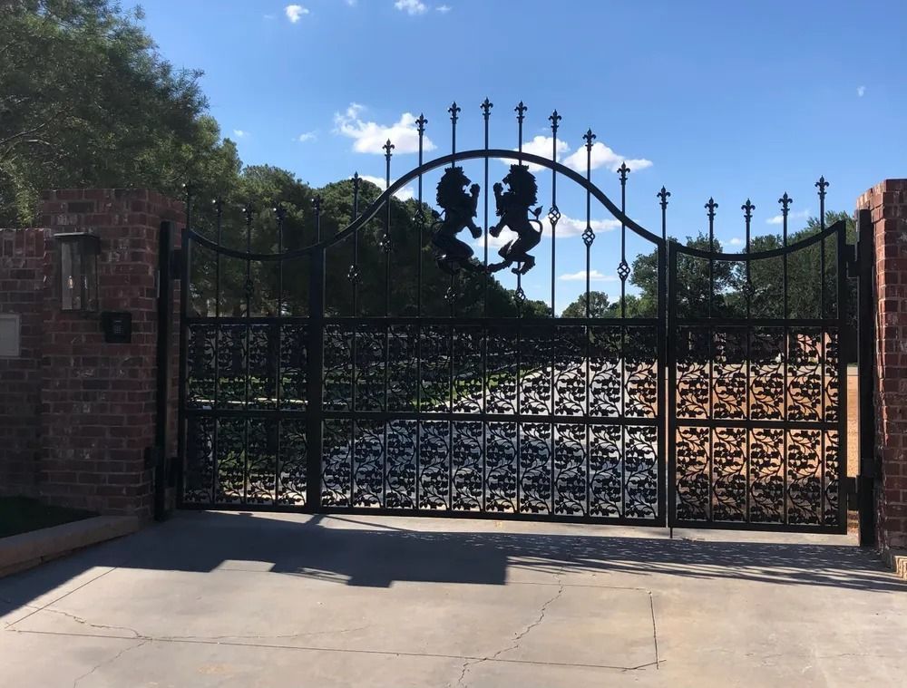 Black iron gates with lion silhouettes, brick pillars, driveway, and blue sky.