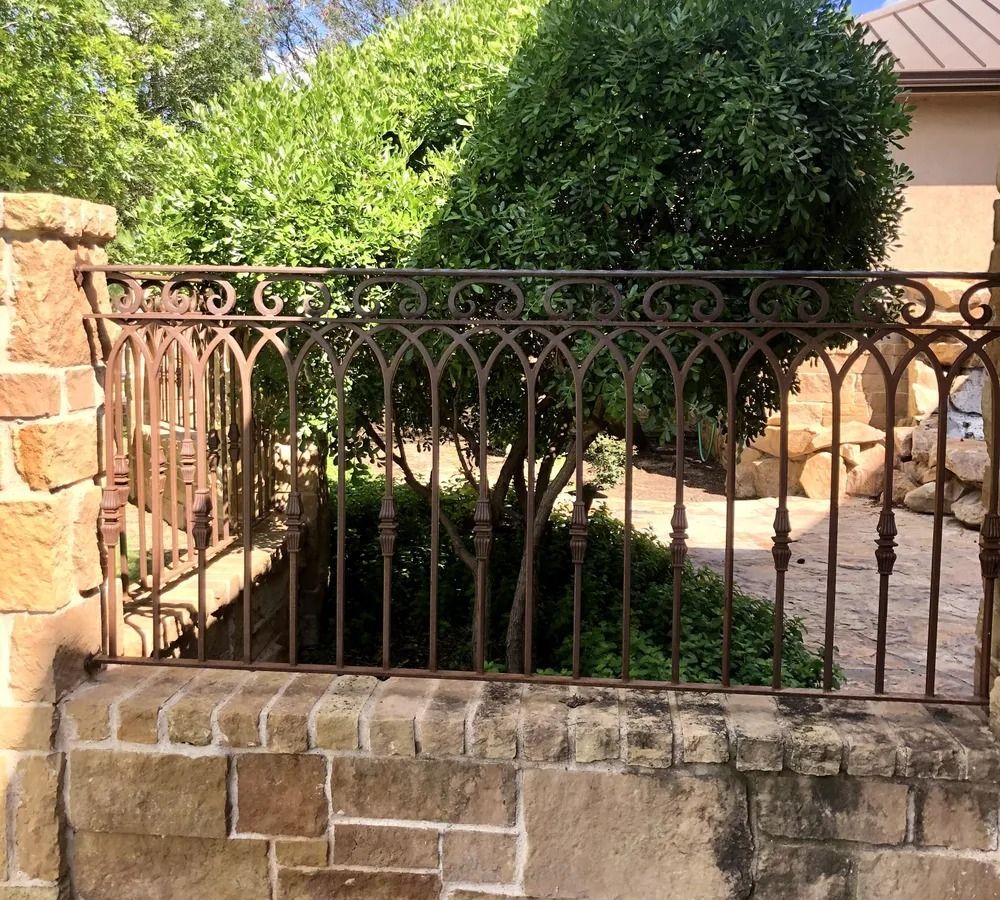 Rusty metal fence atop a stone wall, with greenery in the background.
