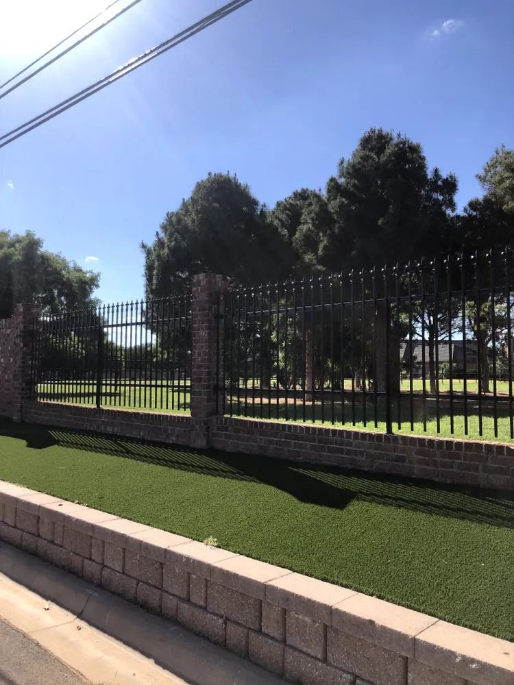 Black wrought iron fence with brick pillars and wall, green turf, trees, and blue sky.