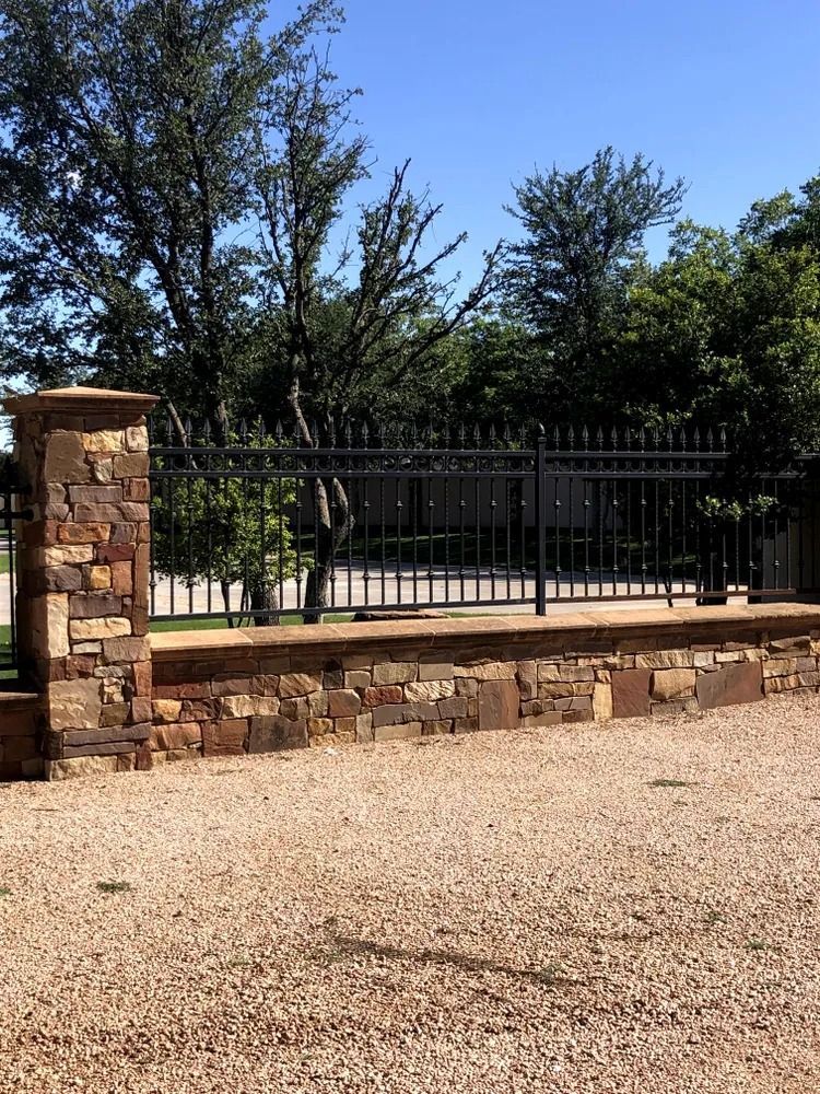 Stone wall with black metal fence, gravel driveway, and trees under a blue sky.