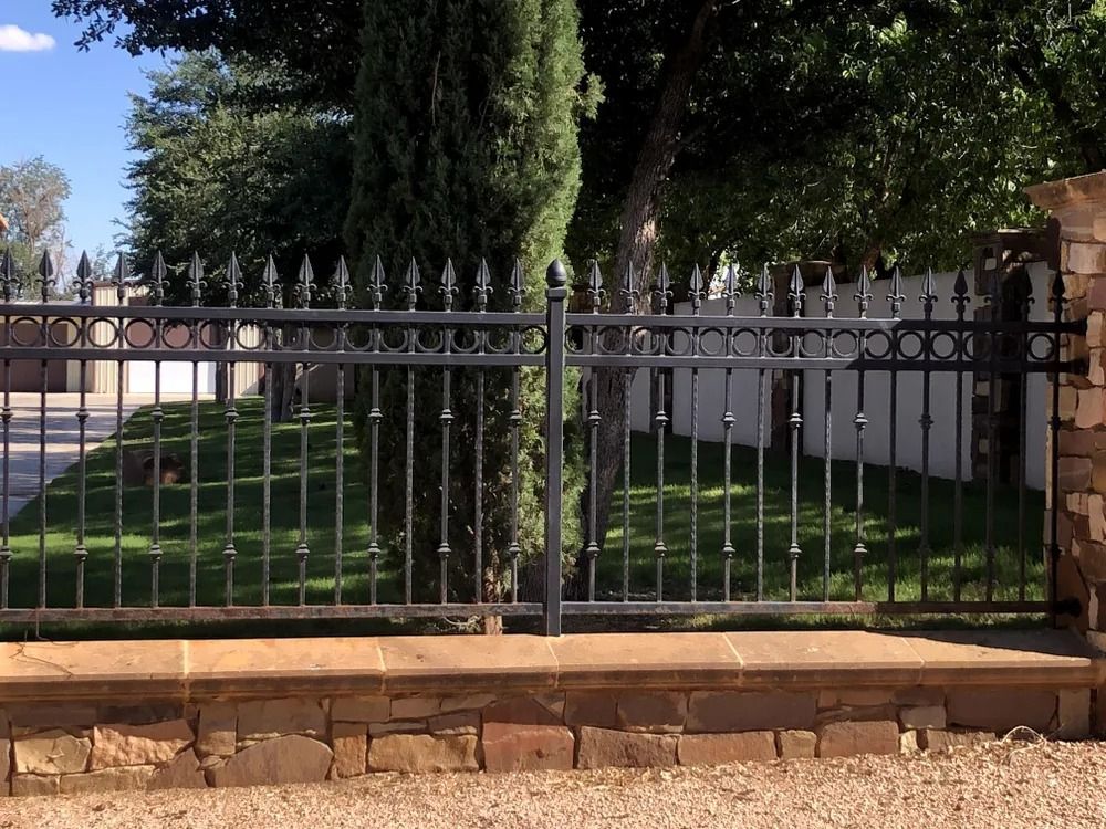 Black metal fence atop a stone wall, in front of grass and trees.