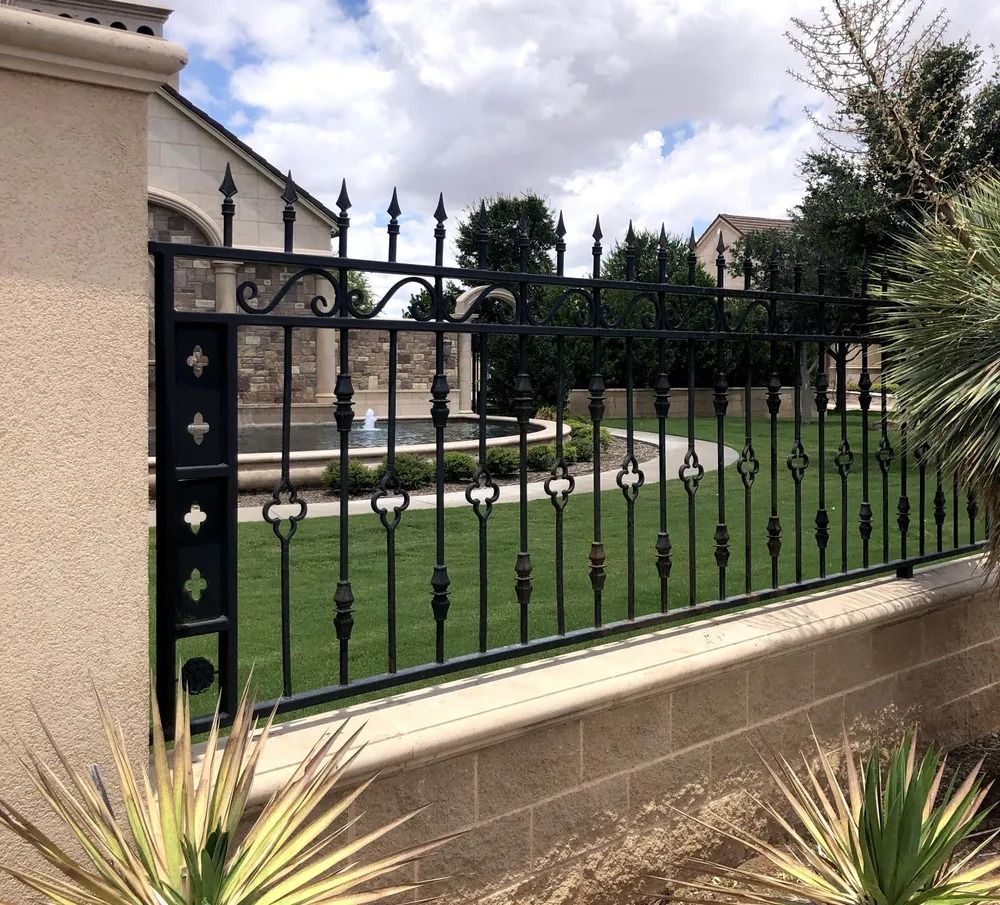 Black wrought iron fence on a low stone wall, with a lawn, trees, and a fountain in the background.