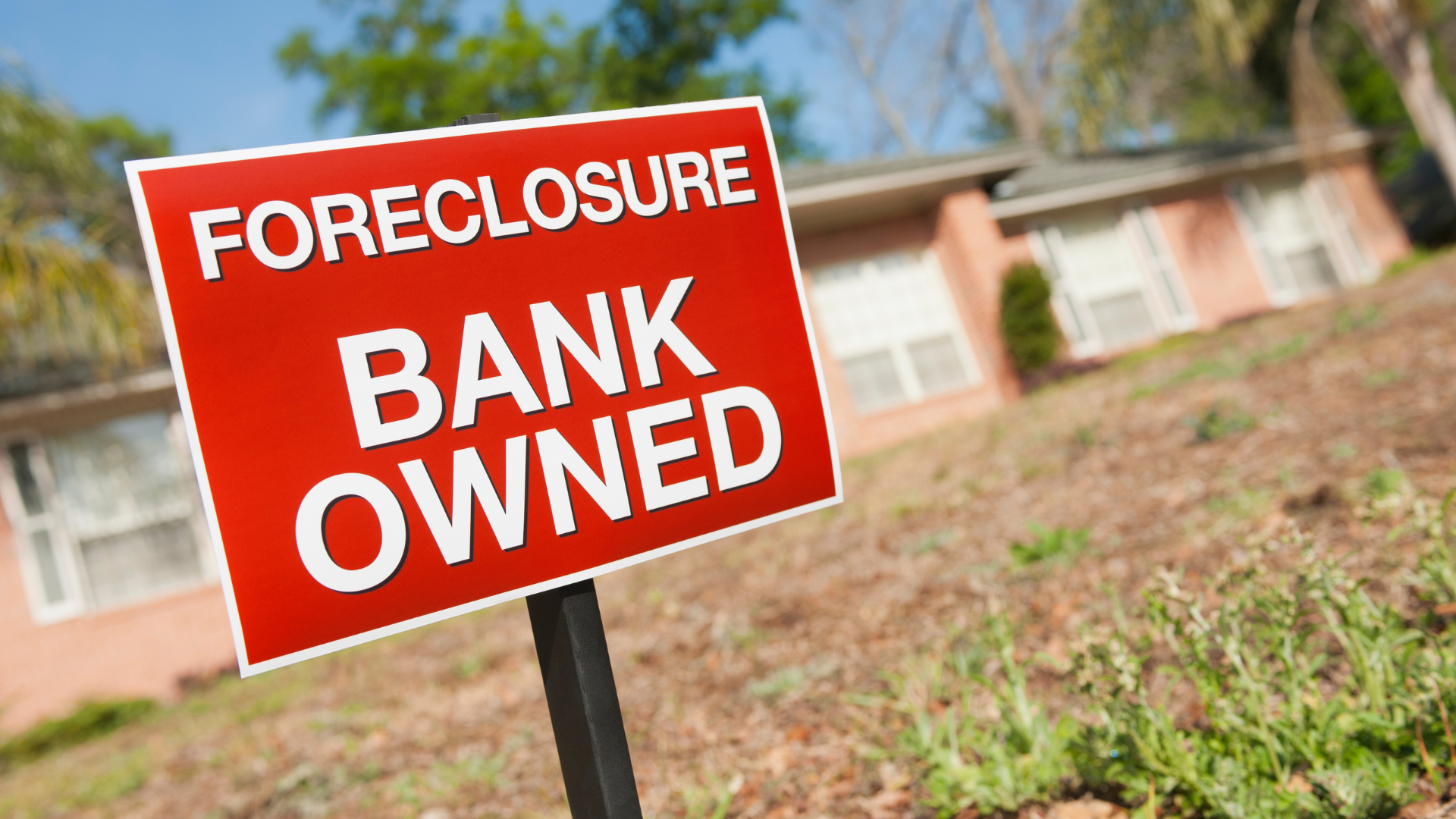 Red foreclosure sign reading “BANK OWNED” in front of a blurred house.