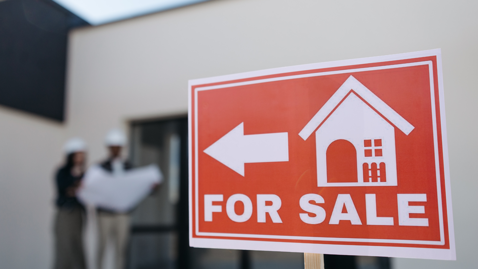 Red “For Sale” sign with white house icon and arrow in front of two blurred people outdoors