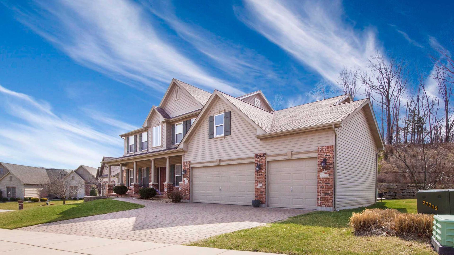 Suburban two-story beige house with a garage, brick accents, and a front porch under a blue sky