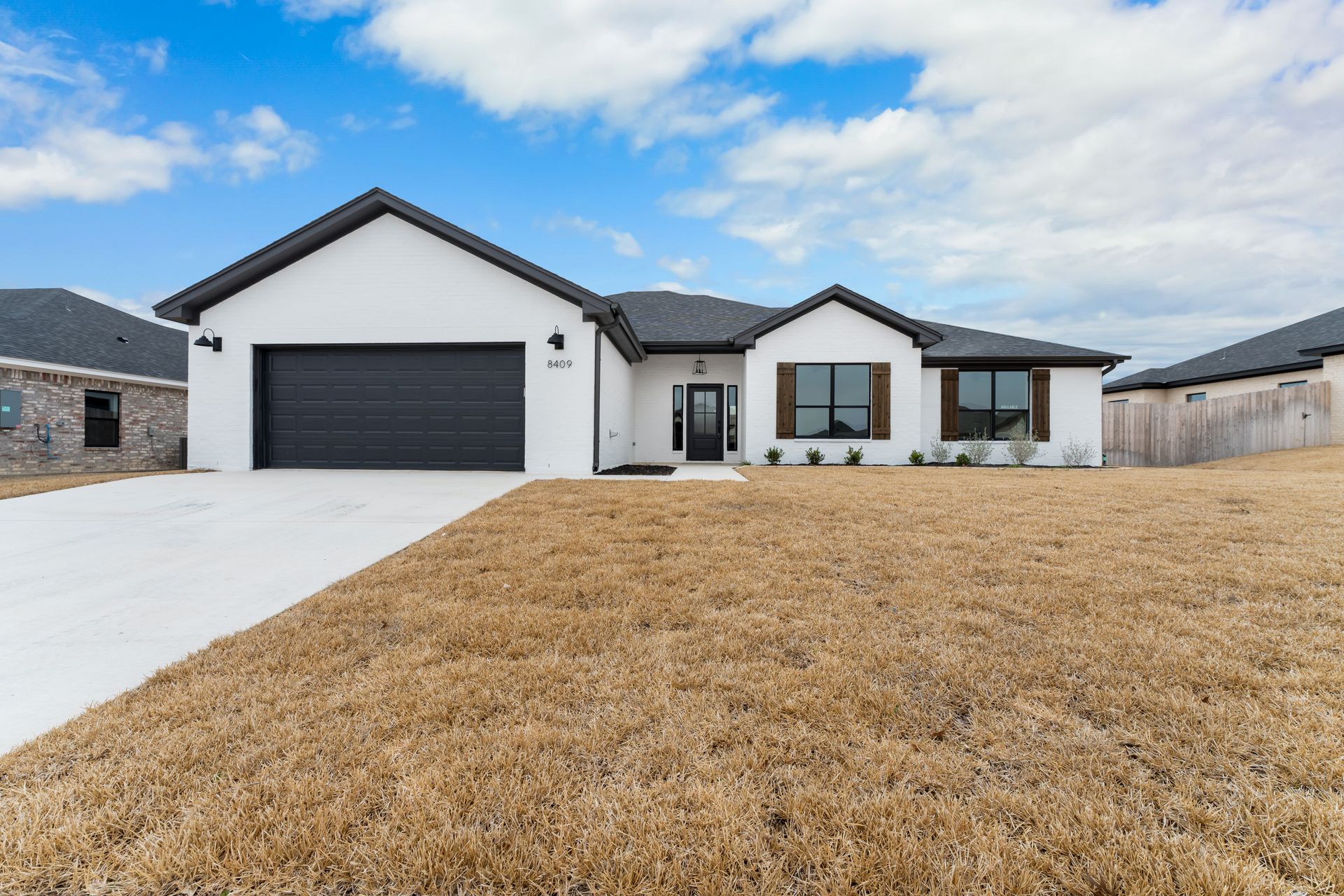 White modern house with black garage door and trim, brown lawn under a blue sky.