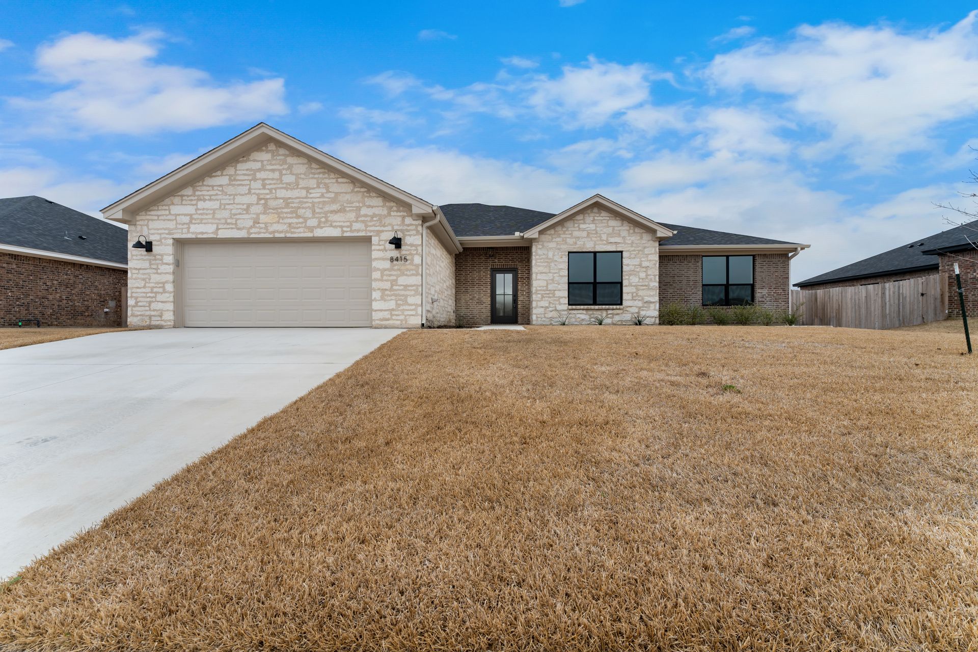 A single-story home with stone facade, beige garage door, and brown lawn under a blue sky.