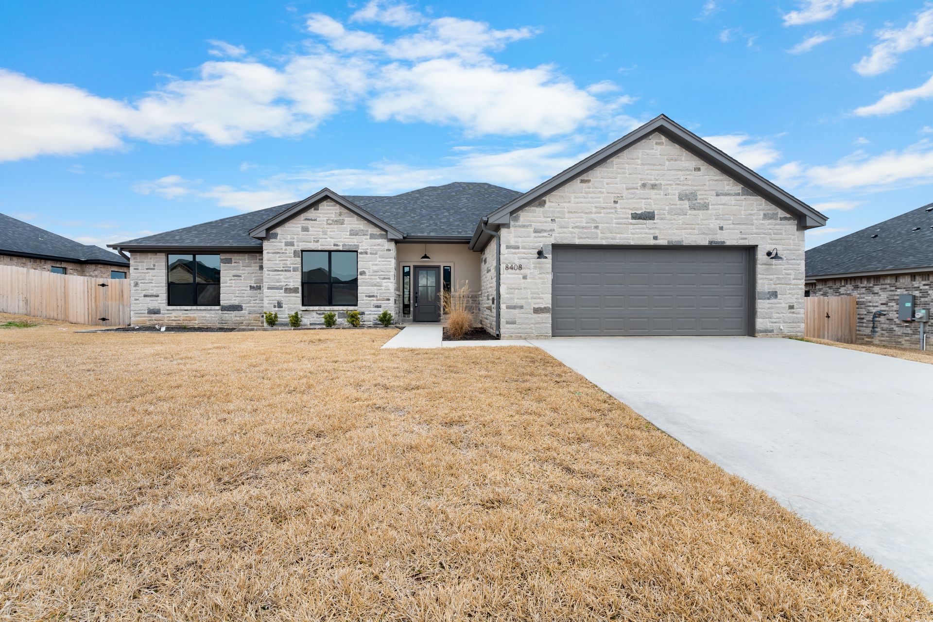 Single-story brick home with gray roof, driveway, and garage door under a blue sky.