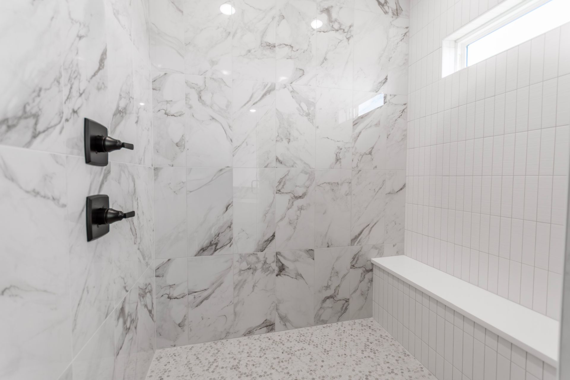 Marble-tiled shower with black fixtures and a white bench. The floor has small, speckled white tiles.