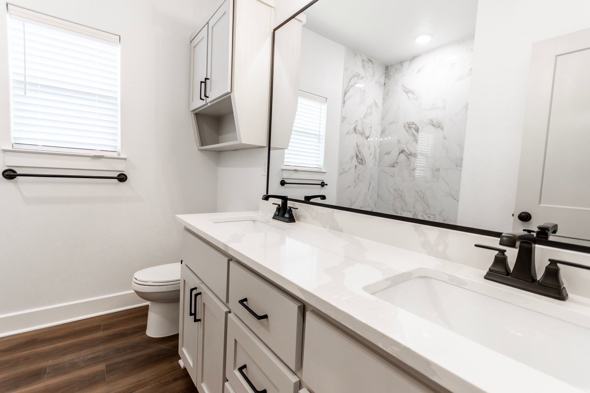Modern white bathroom with double sink vanity, toilet, shower, and window. Black fixtures and trim.