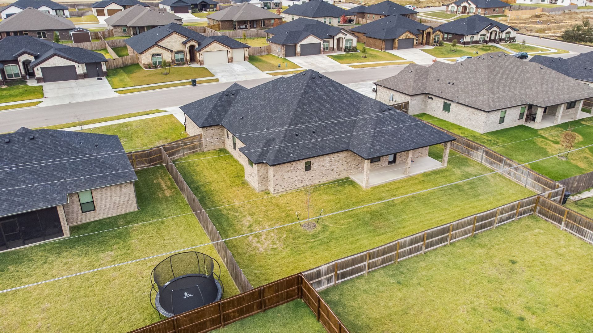 Aerial view of suburban houses with dark roofs and green lawns, some fenced.