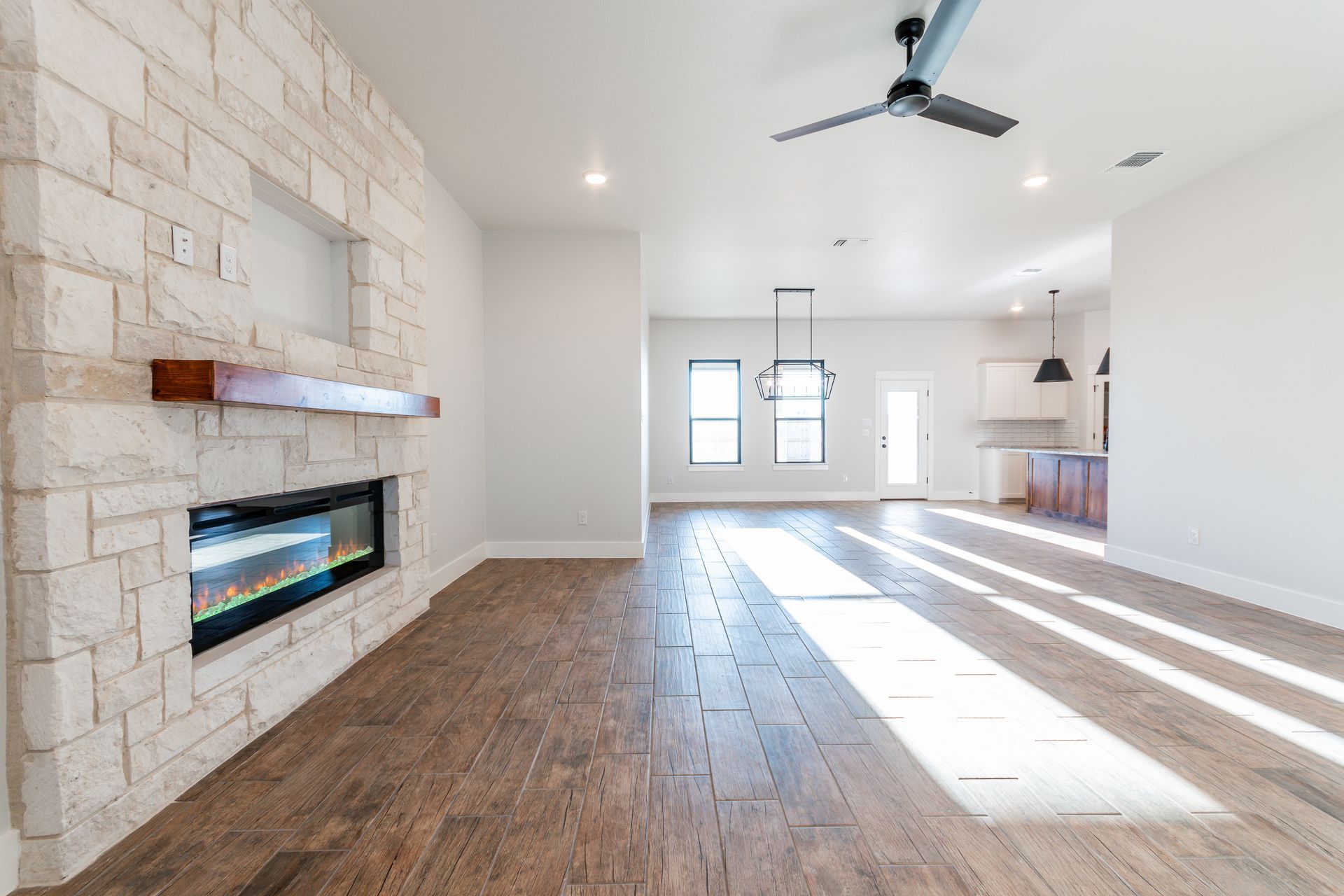 Living room with stone fireplace, wooden floor, and a ceiling fan. Bright sunlight streams in.