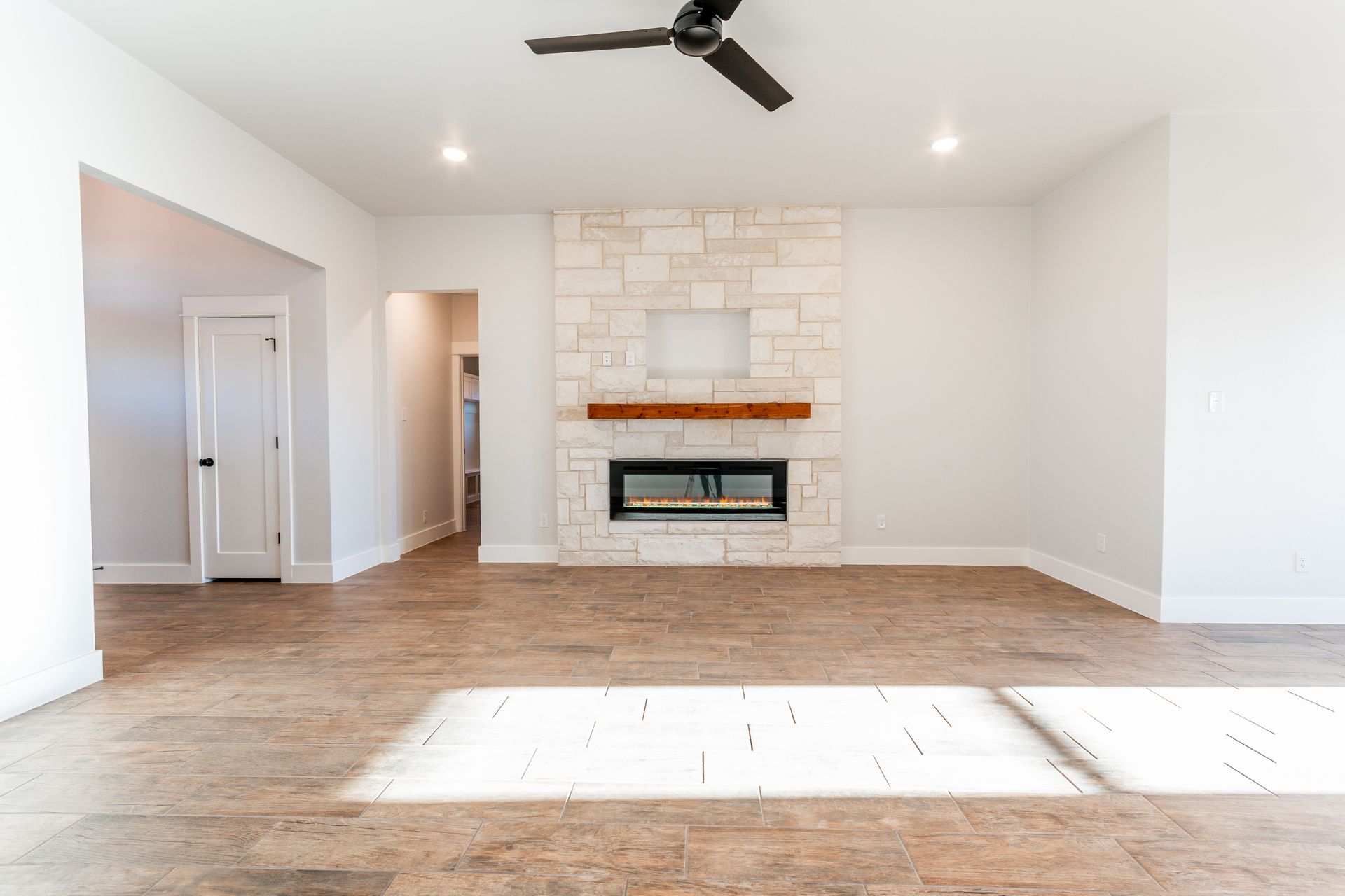Empty living room with light-colored flooring, white walls, stone fireplace, and a ceiling fan.