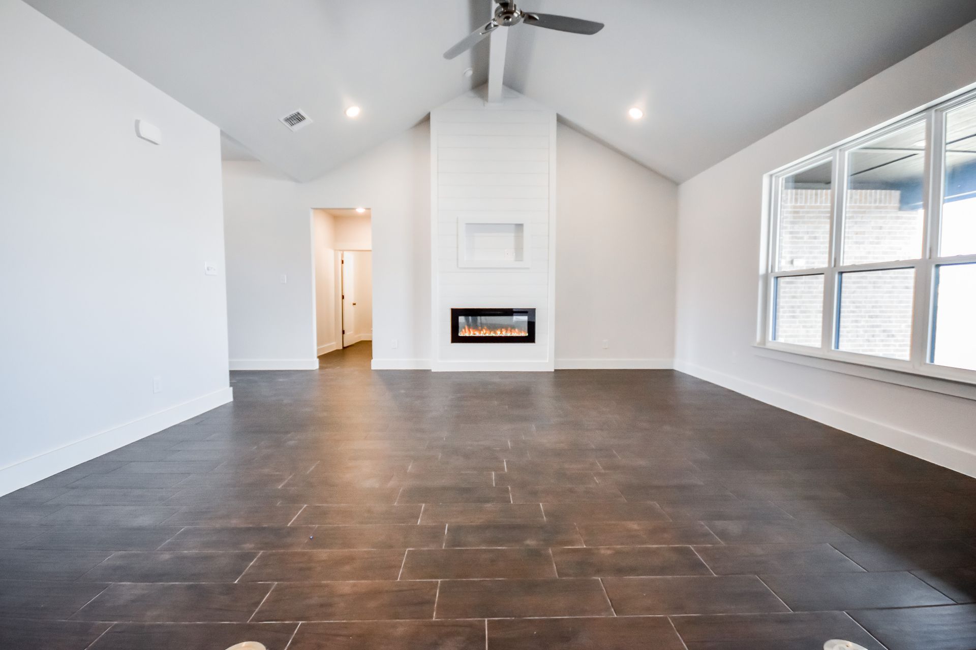 Empty, modern living room with fireplace, high ceiling, large window. Dark tile floor, white walls.
