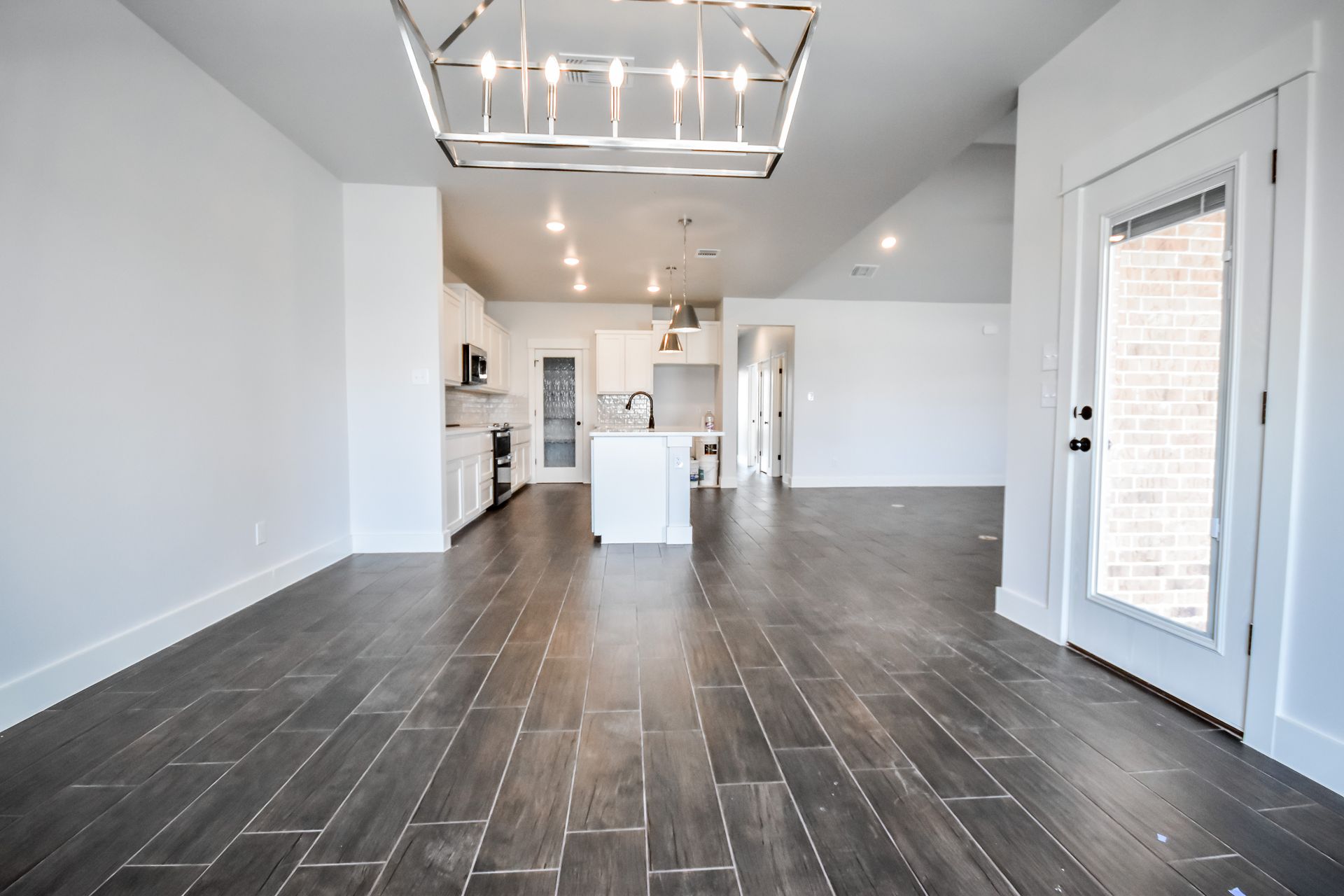 Empty interior with dark wood-look floor, white walls, and kitchen in the background. Modern light fixture hangs from the ceiling.