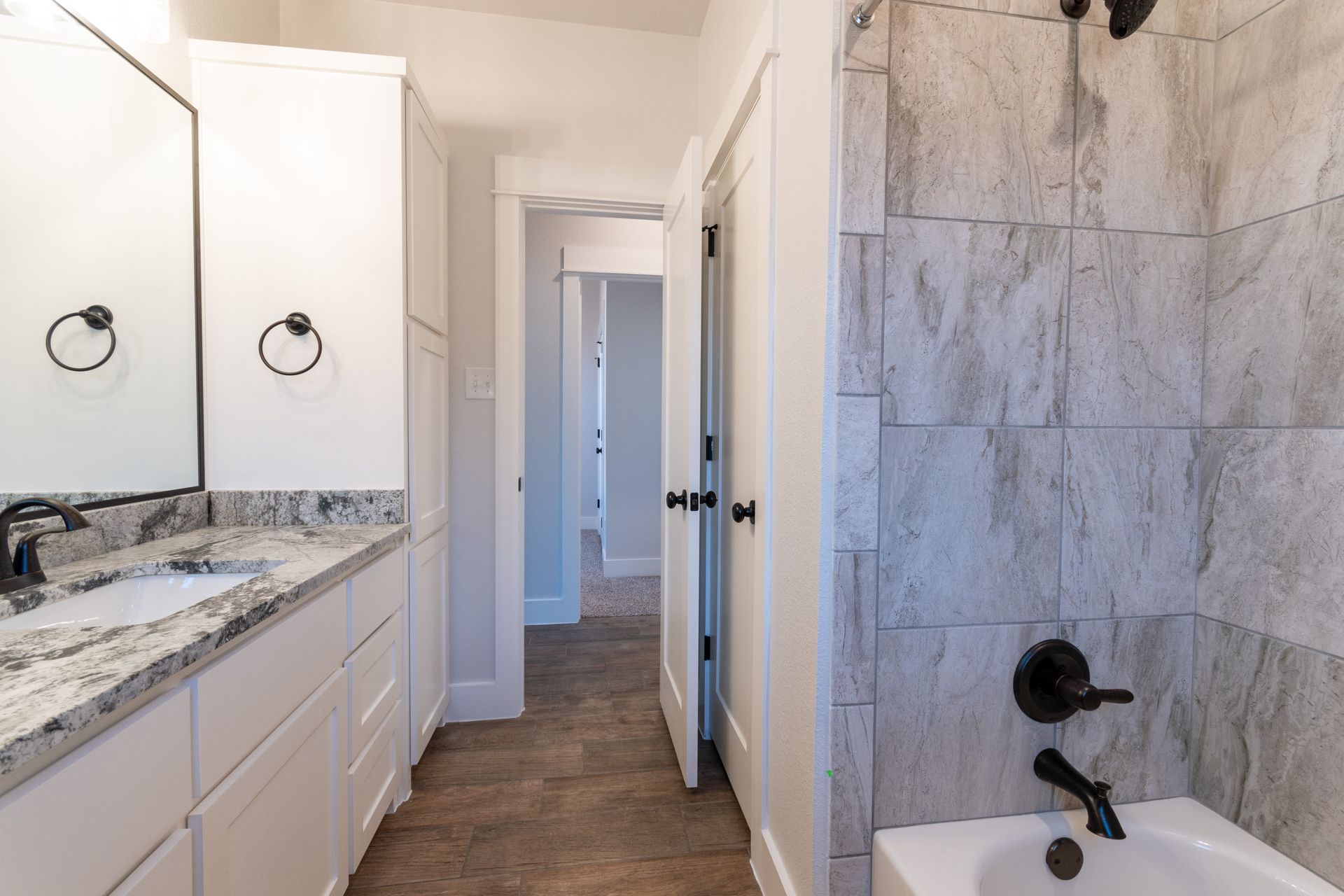 Bathroom with white cabinets, gray granite countertop, and gray tiled shower. Doorway leads to a hallway.