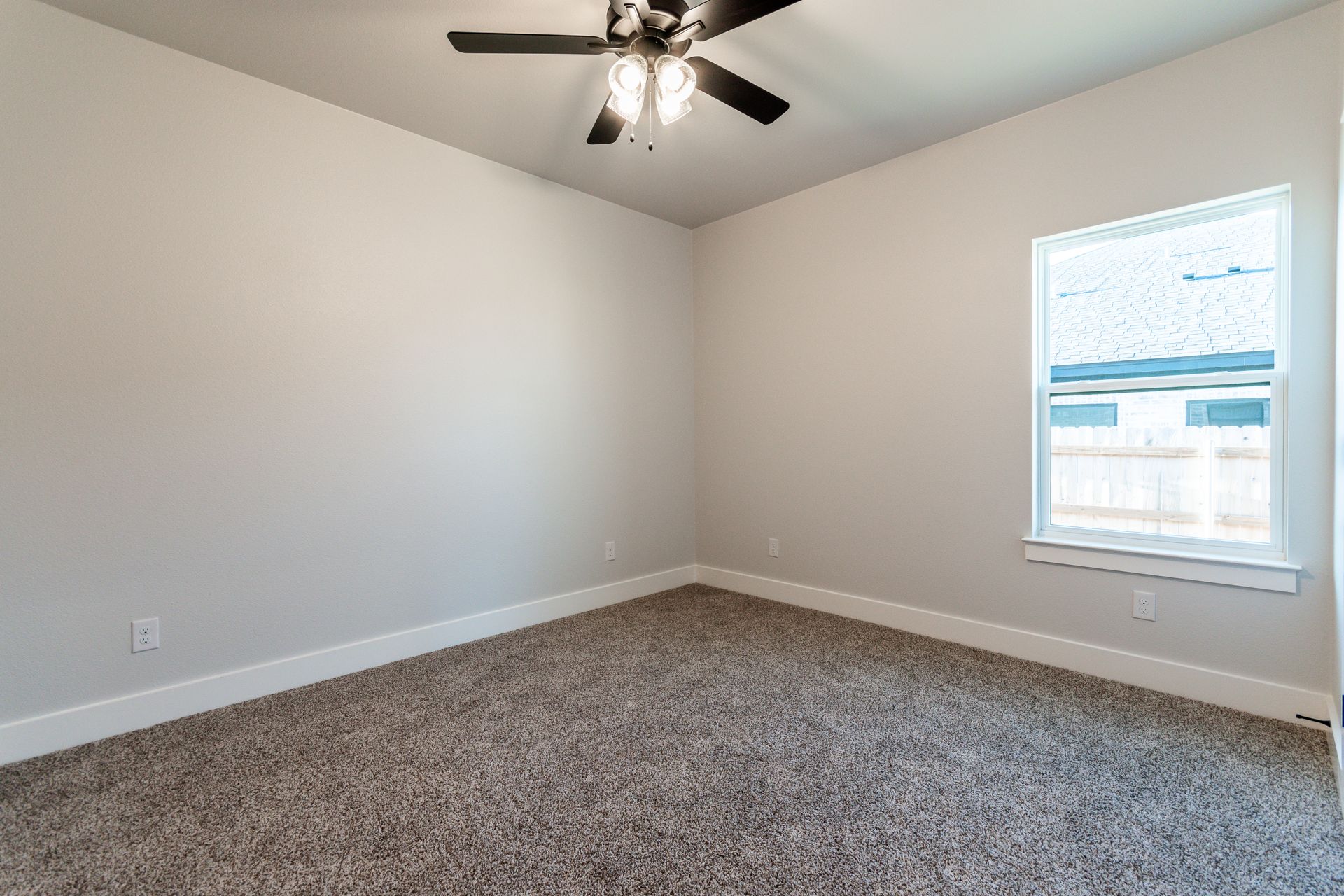 Empty bedroom with beige carpet, light gray walls, and a ceiling fan. A window lets in sunlight.
