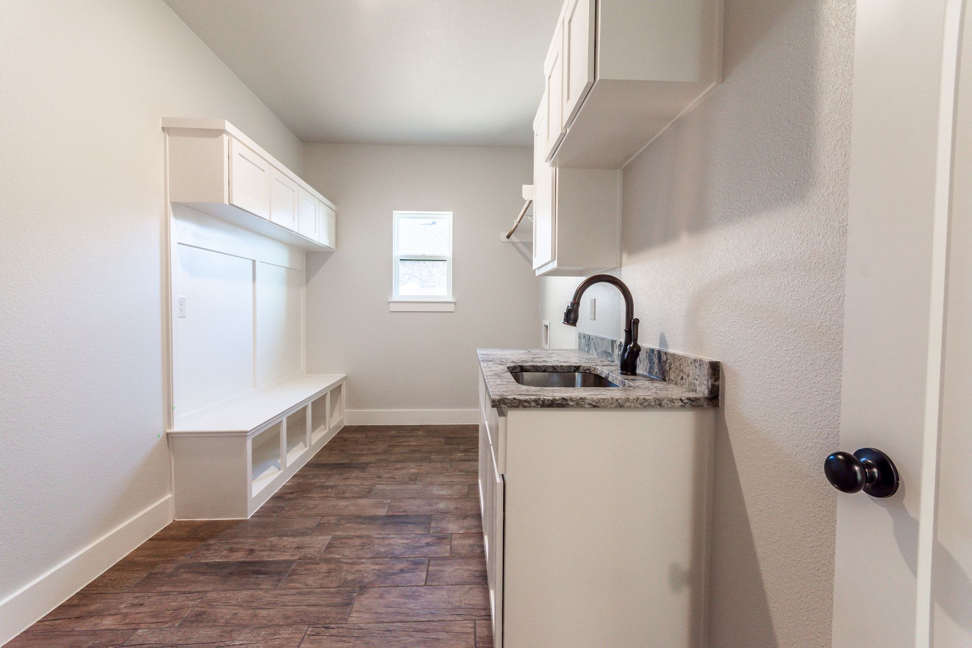 Laundry room with white cabinets, bench, sink, and dark faucet against a textured wall and wood-look floor.