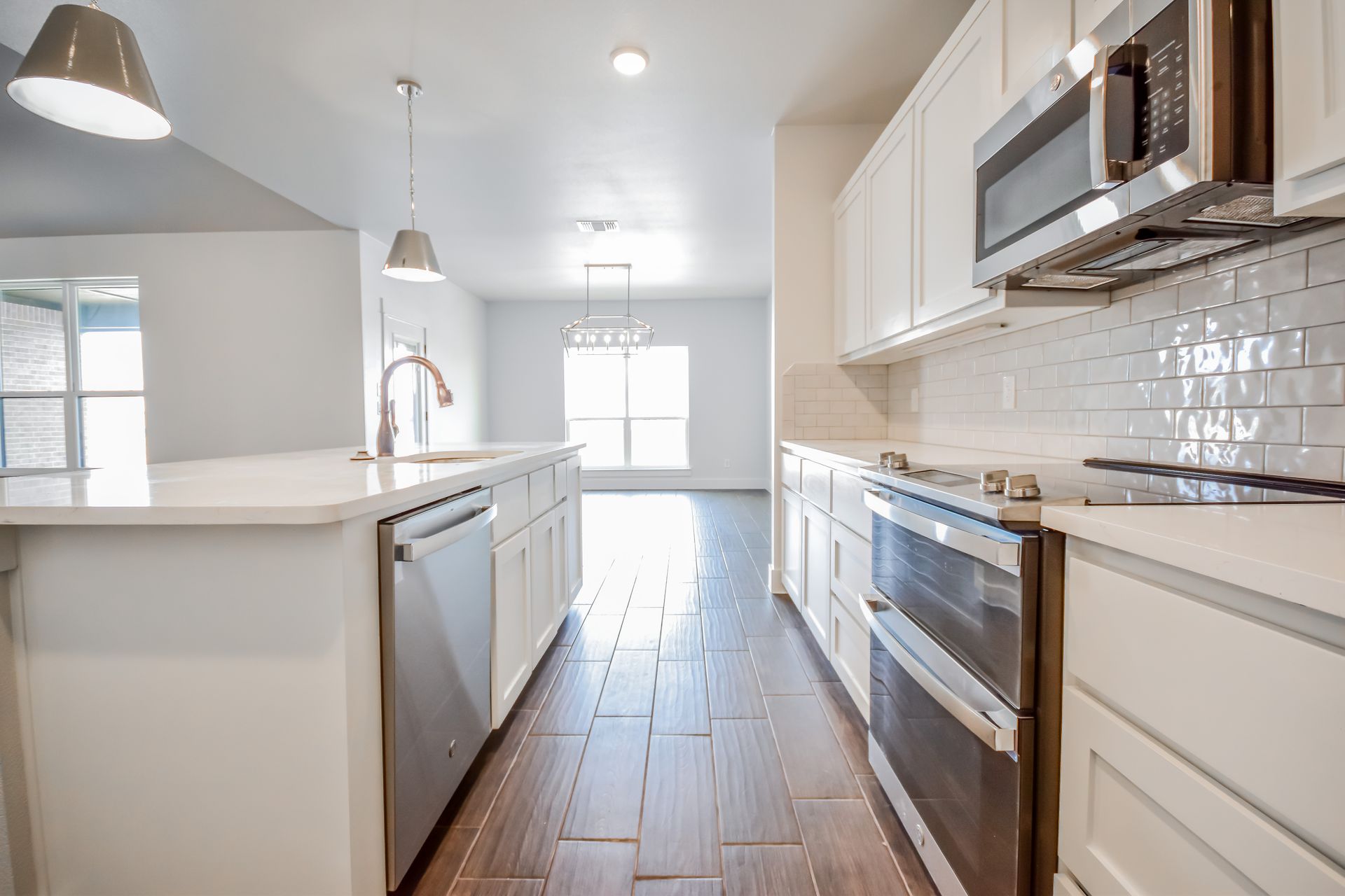 Bright kitchen with white cabinets, island, and appliances; a view into a living space with natural light.