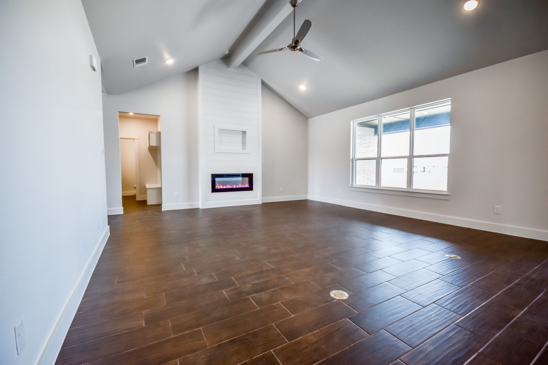 Spacious, empty room with brown tile flooring, fireplace, and large window. White walls and vaulted ceiling.
