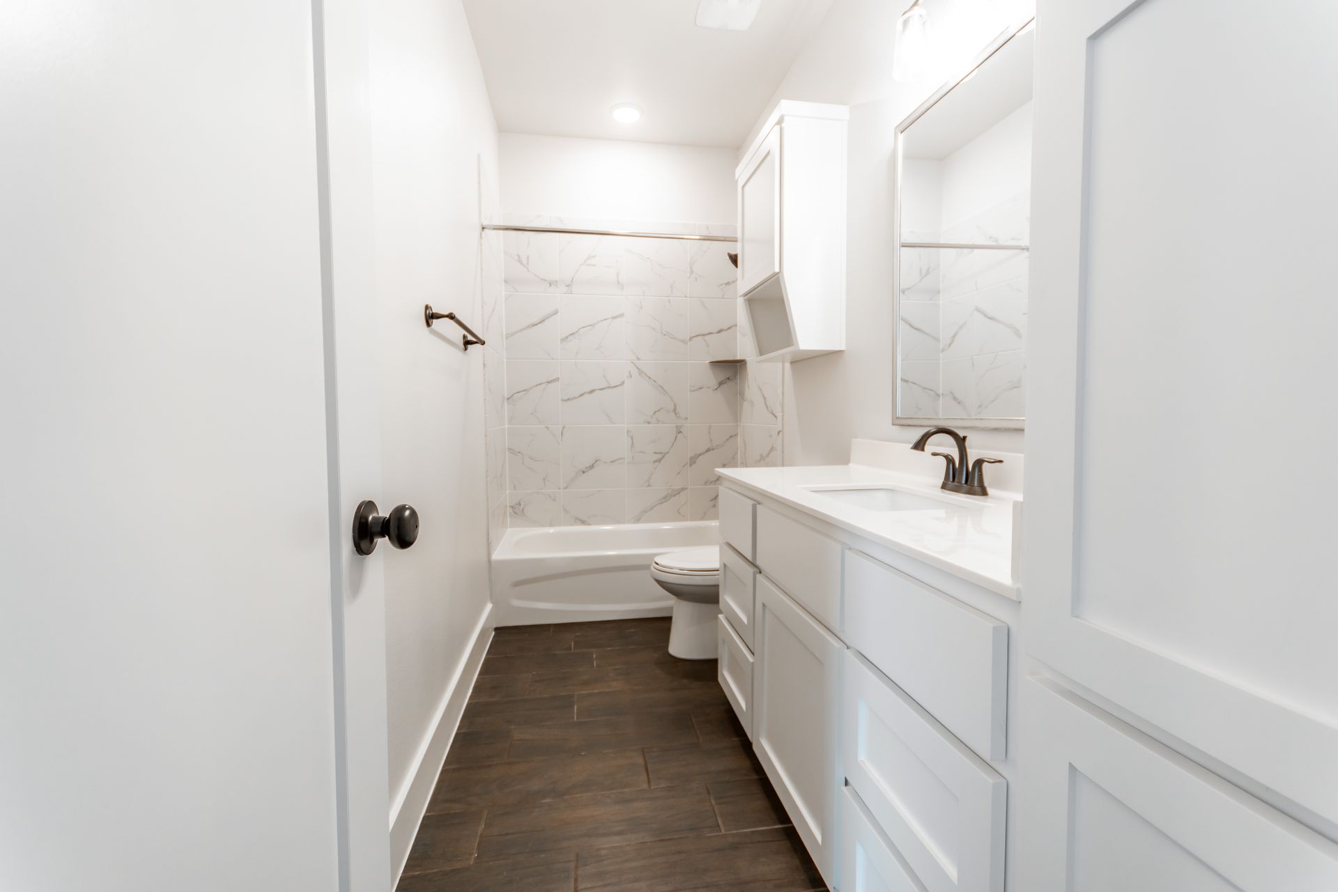 White bathroom with dark tile floor, white vanity and toilet, marble tile shower, black hardware.