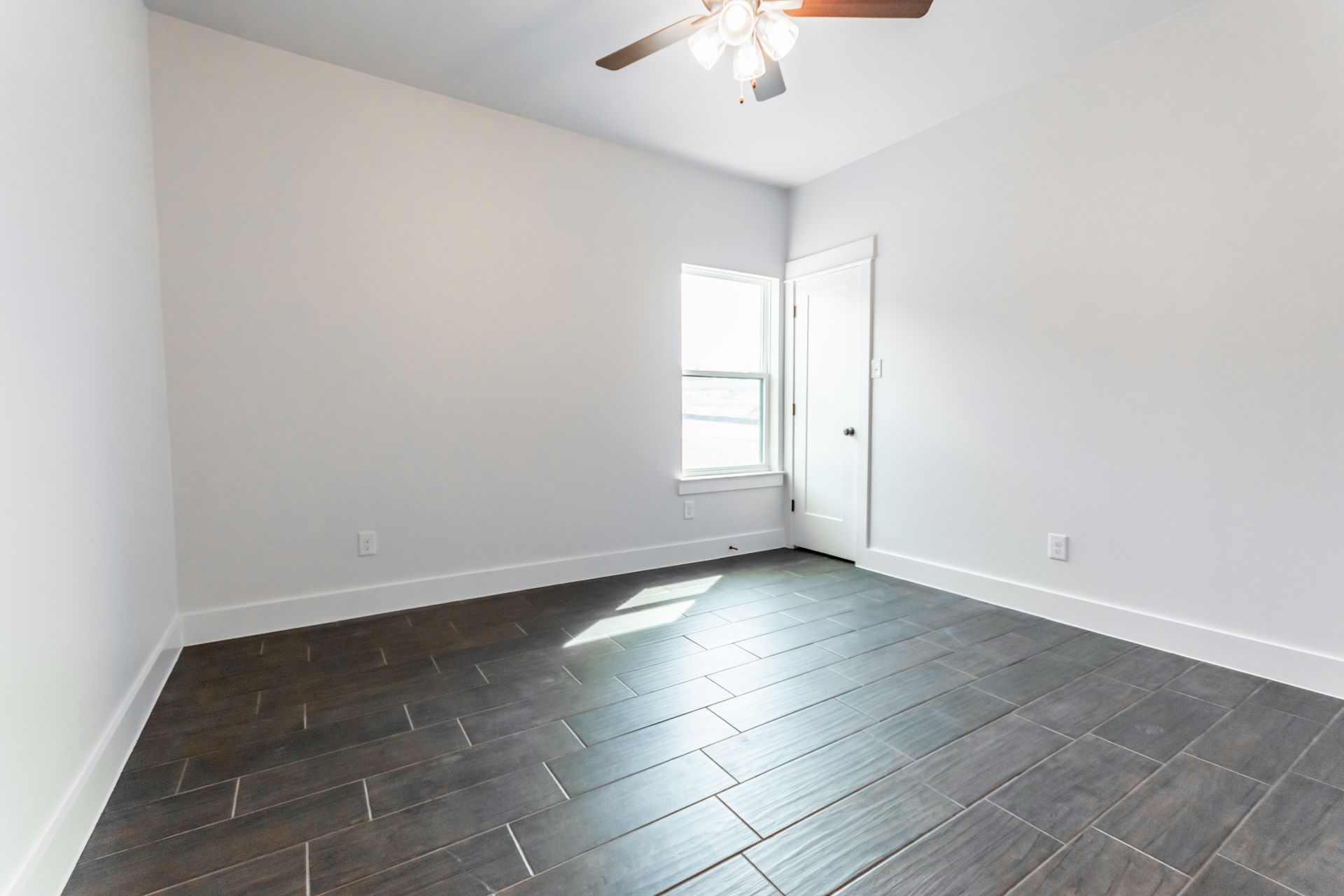 Empty bedroom with gray tile floor, white walls, window, door, and ceiling fan.