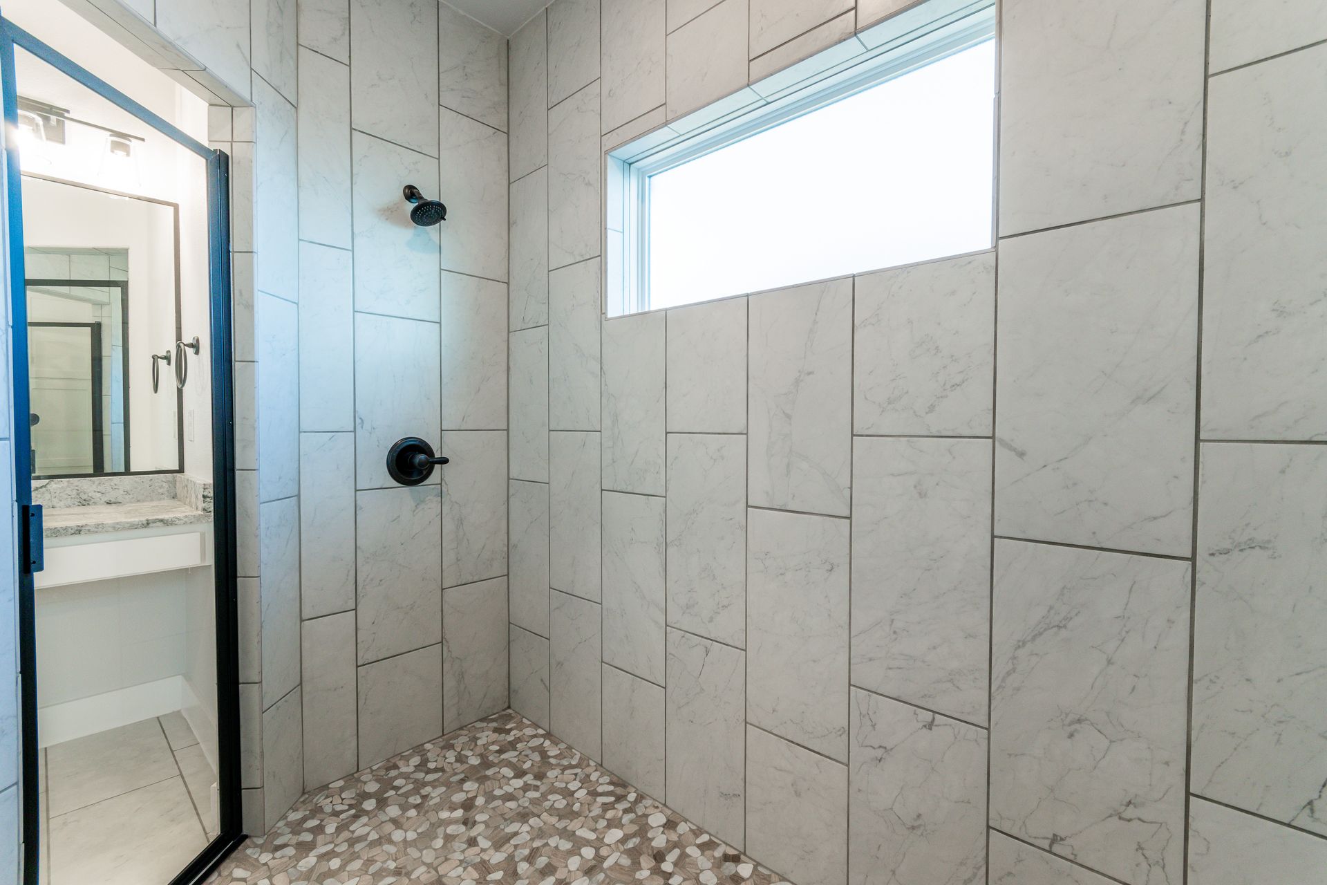 Modern shower with white tiled walls, pebble floor, and black fixtures.
