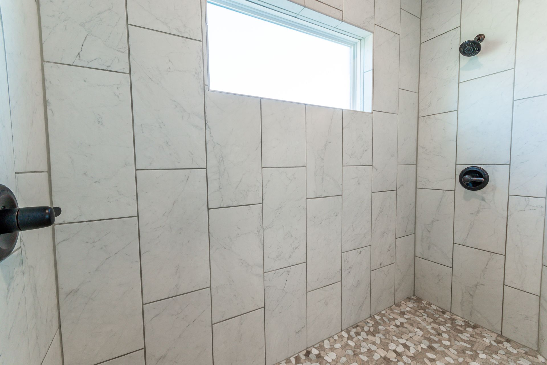 Shower with white tiled walls, pebble floor, and black fixtures.