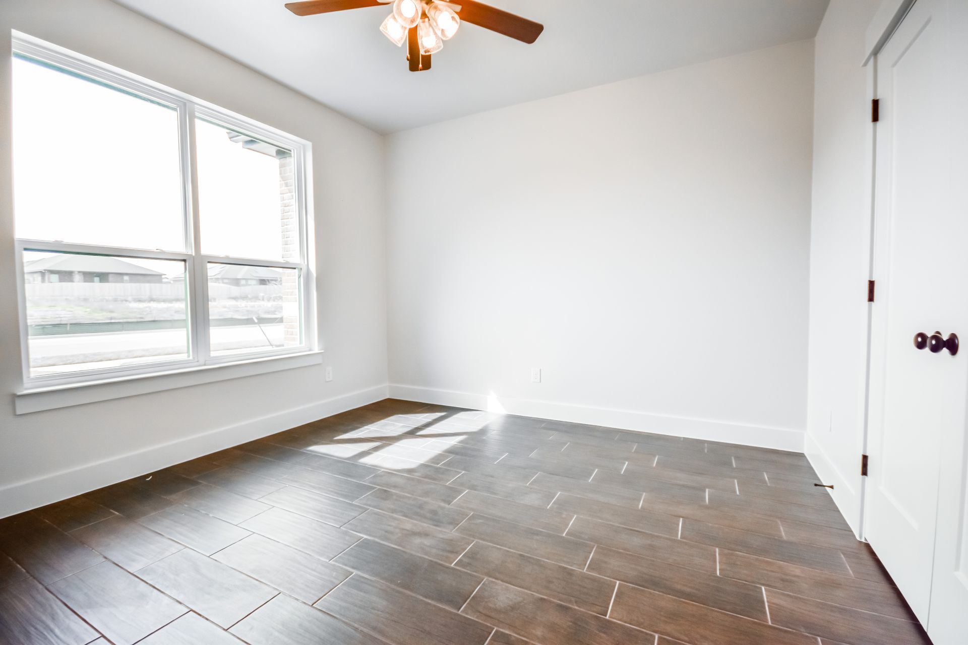 Empty room with wood-look tile flooring, white walls, large window, and ceiling fan.