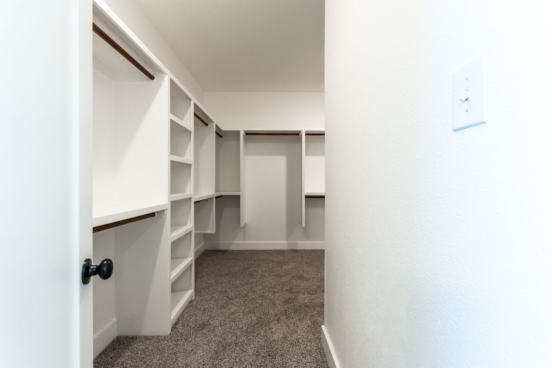 Walk-in closet with white shelving and brown carpet. Door open, light switch visible on the wall.