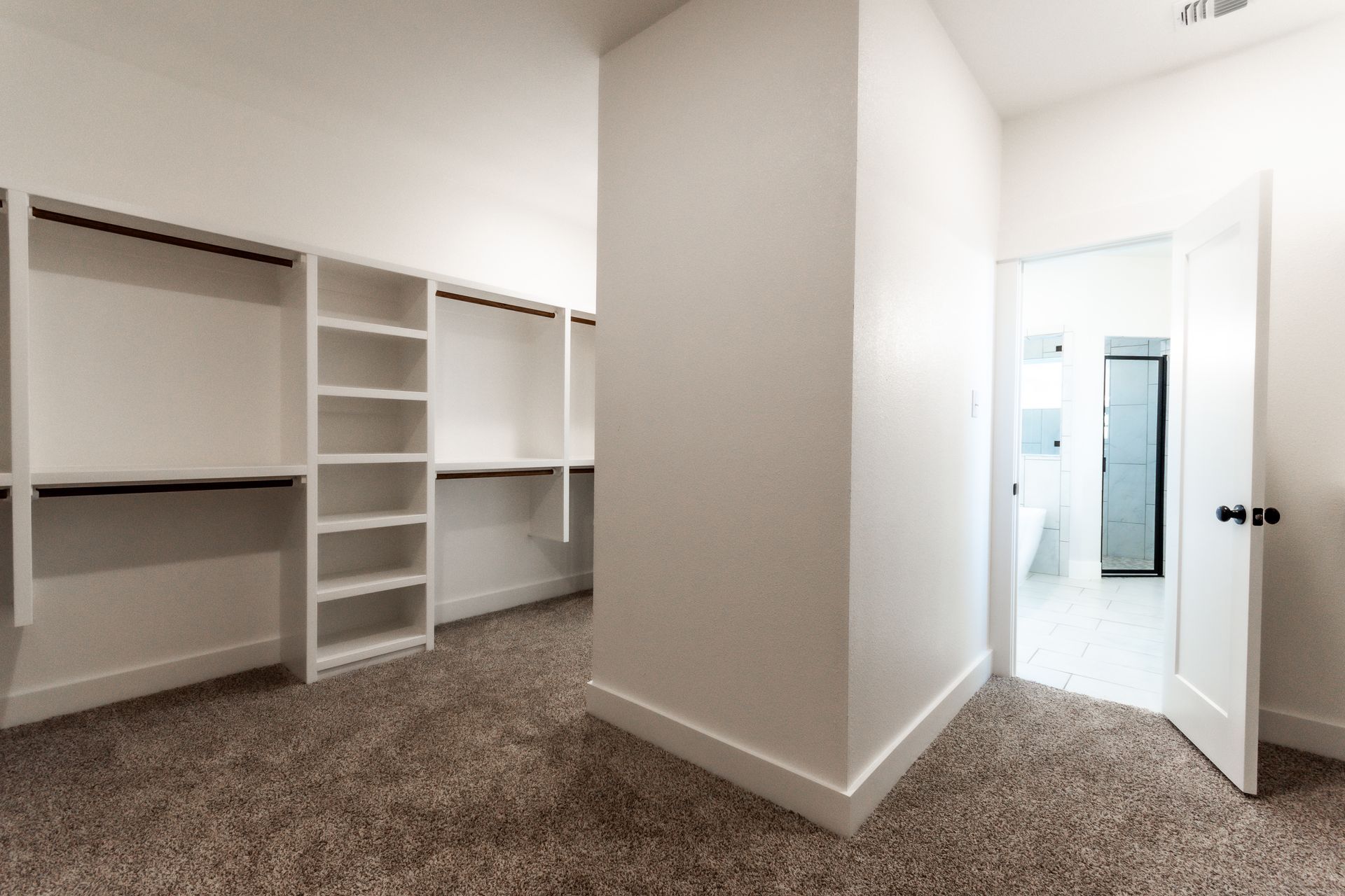 Empty walk-in closet with built-in shelves and rods, carpet floor, doorway leading to a bathroom.