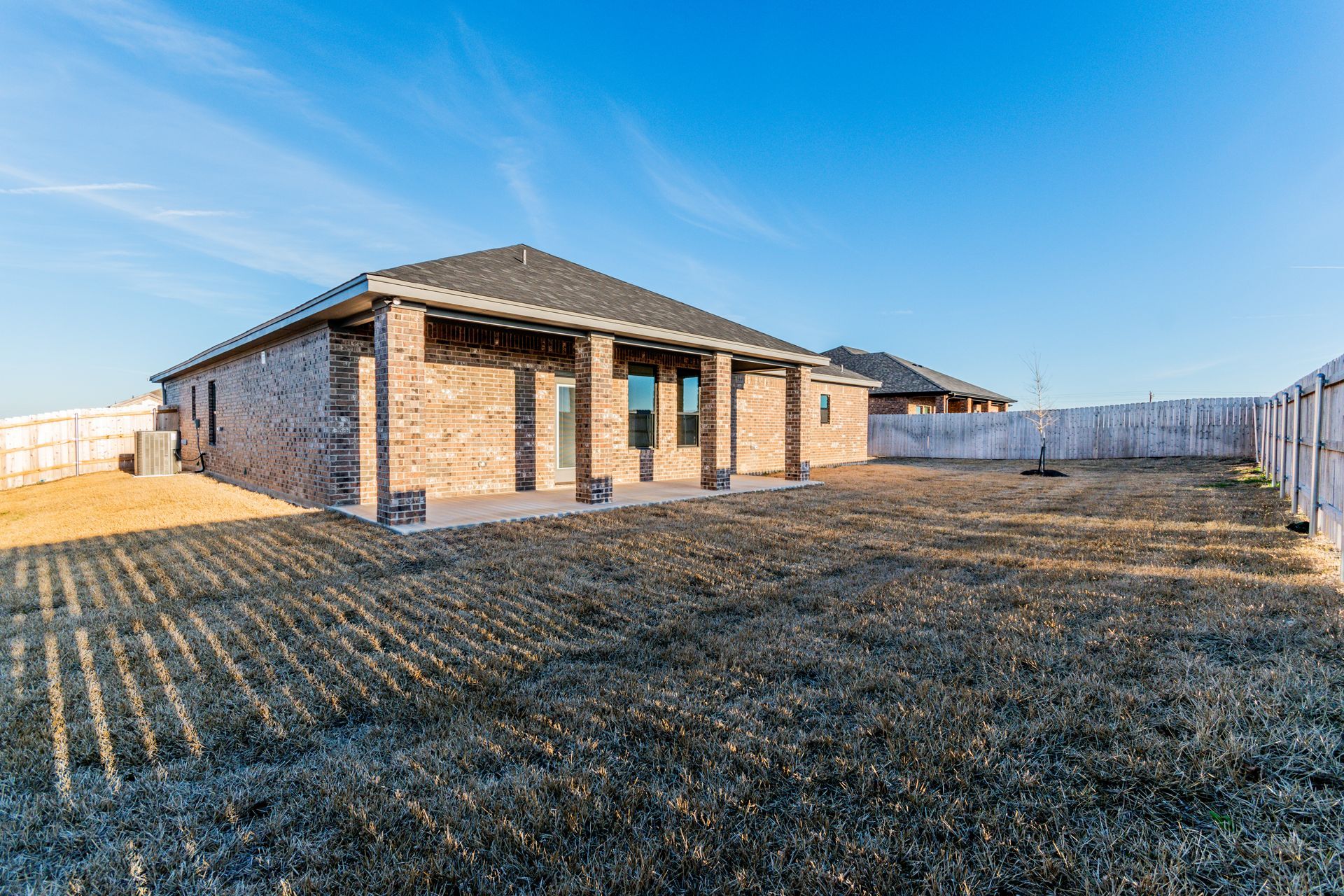 Backyard of a brick home with brown grass, wooden fence, and blue sky.
