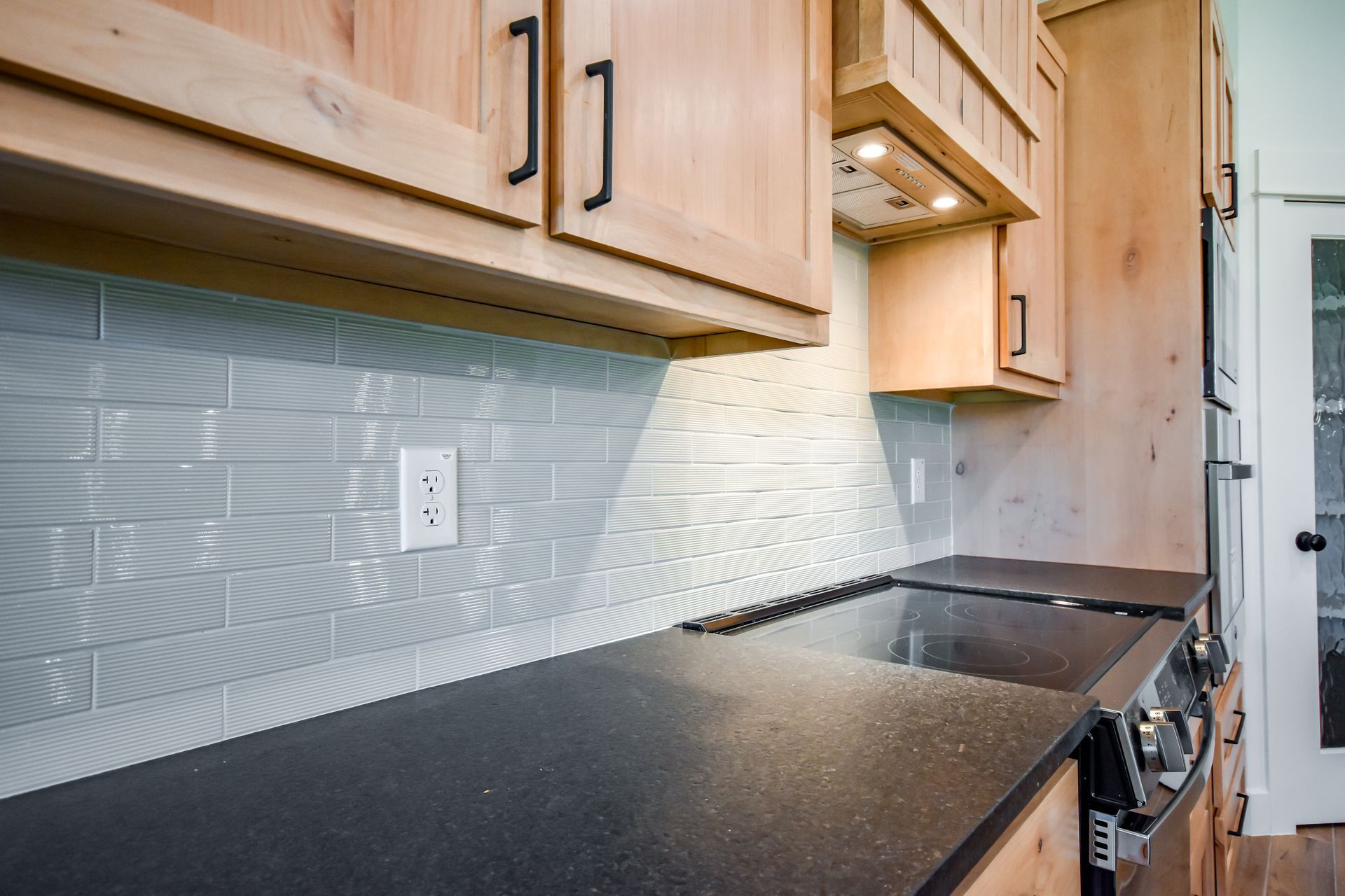 Kitchen with light wood cabinets, gray backsplash, black countertop, and cooktop.