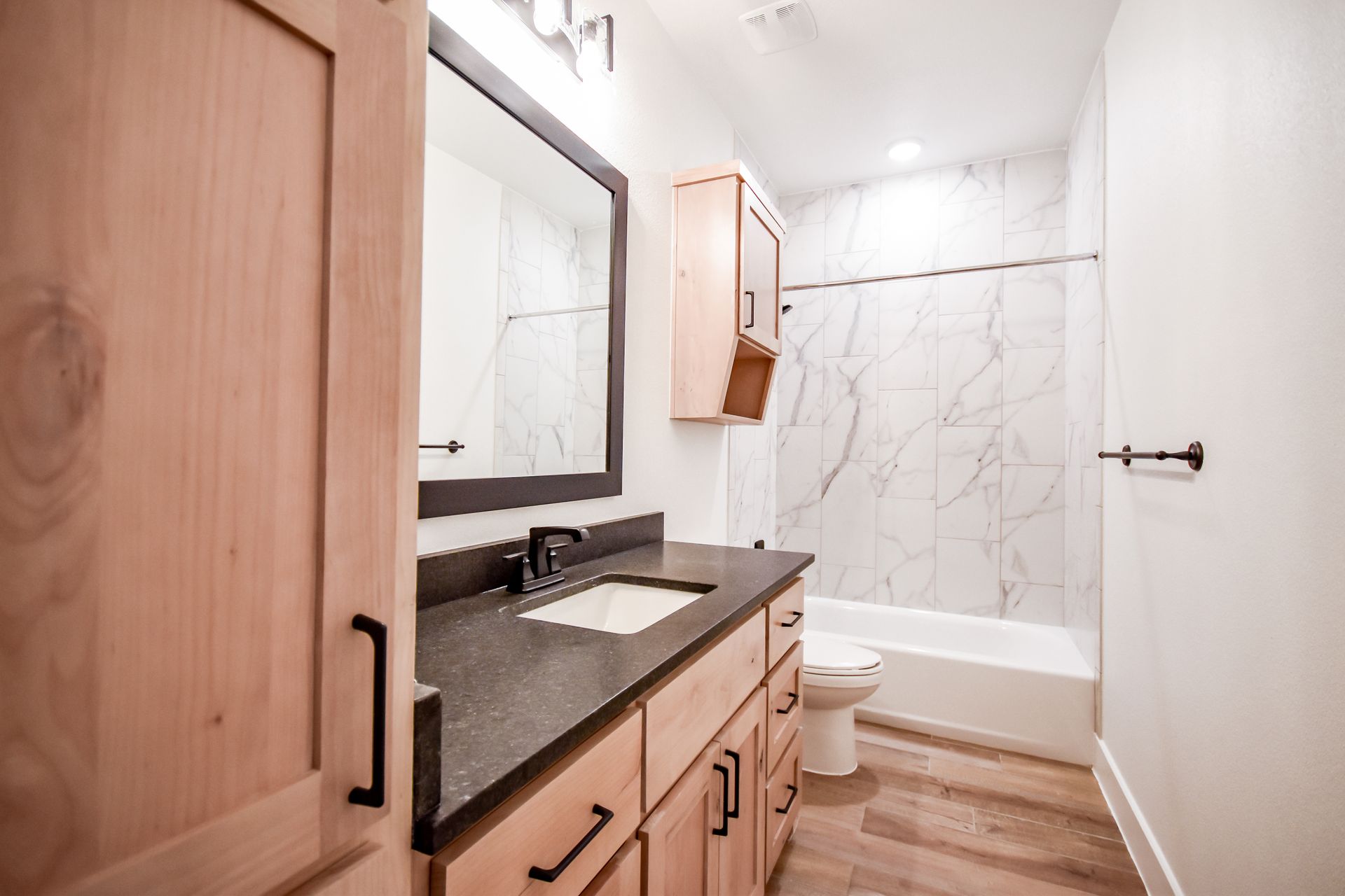 Bathroom with light wood cabinets, dark countertop, marble tiled shower, and a large mirror.
