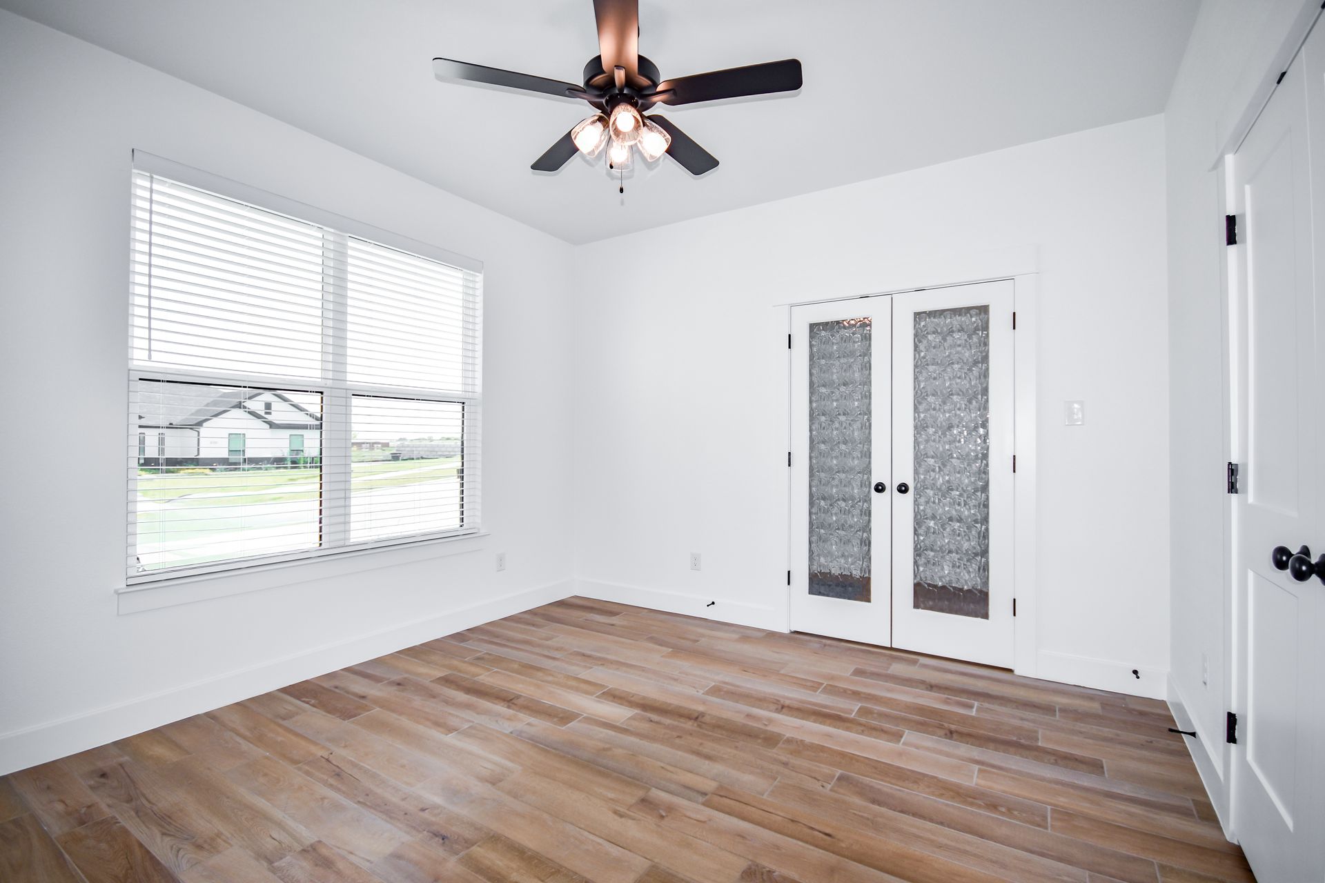Empty room with wood floors, a window with blinds, and French doors. A ceiling fan hangs from the ceiling.