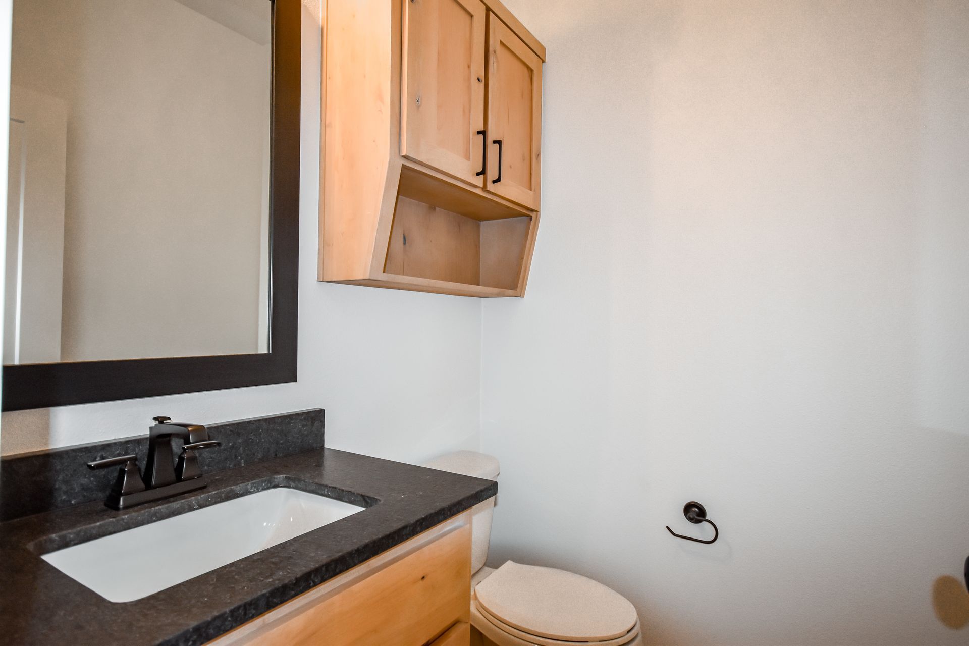Bathroom with vanity, mirror, toilet, and wooden cabinet. White walls, black countertop and faucet.
