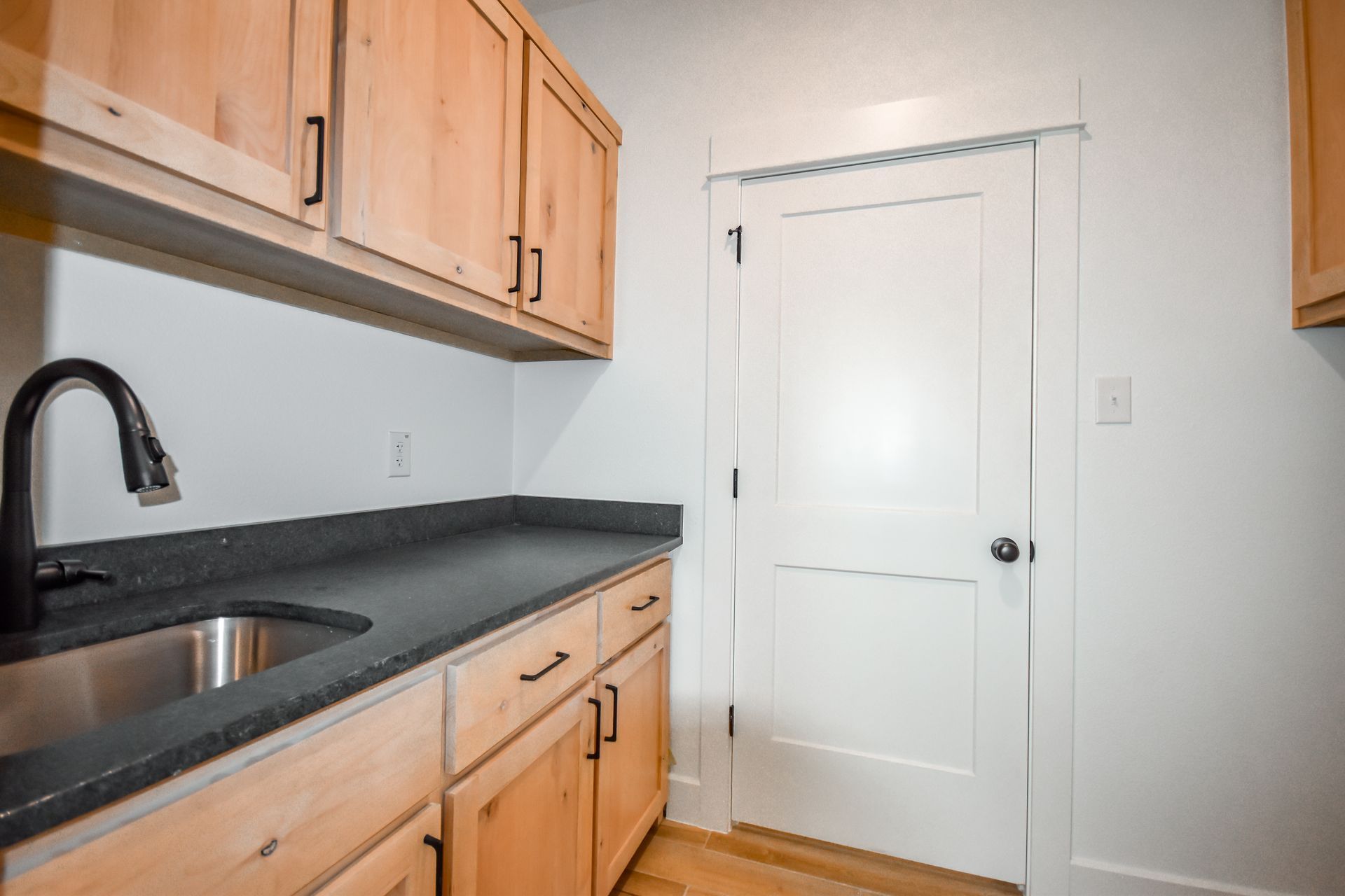 Kitchen with light wood cabinets, dark countertop, stainless steel sink, and white door.