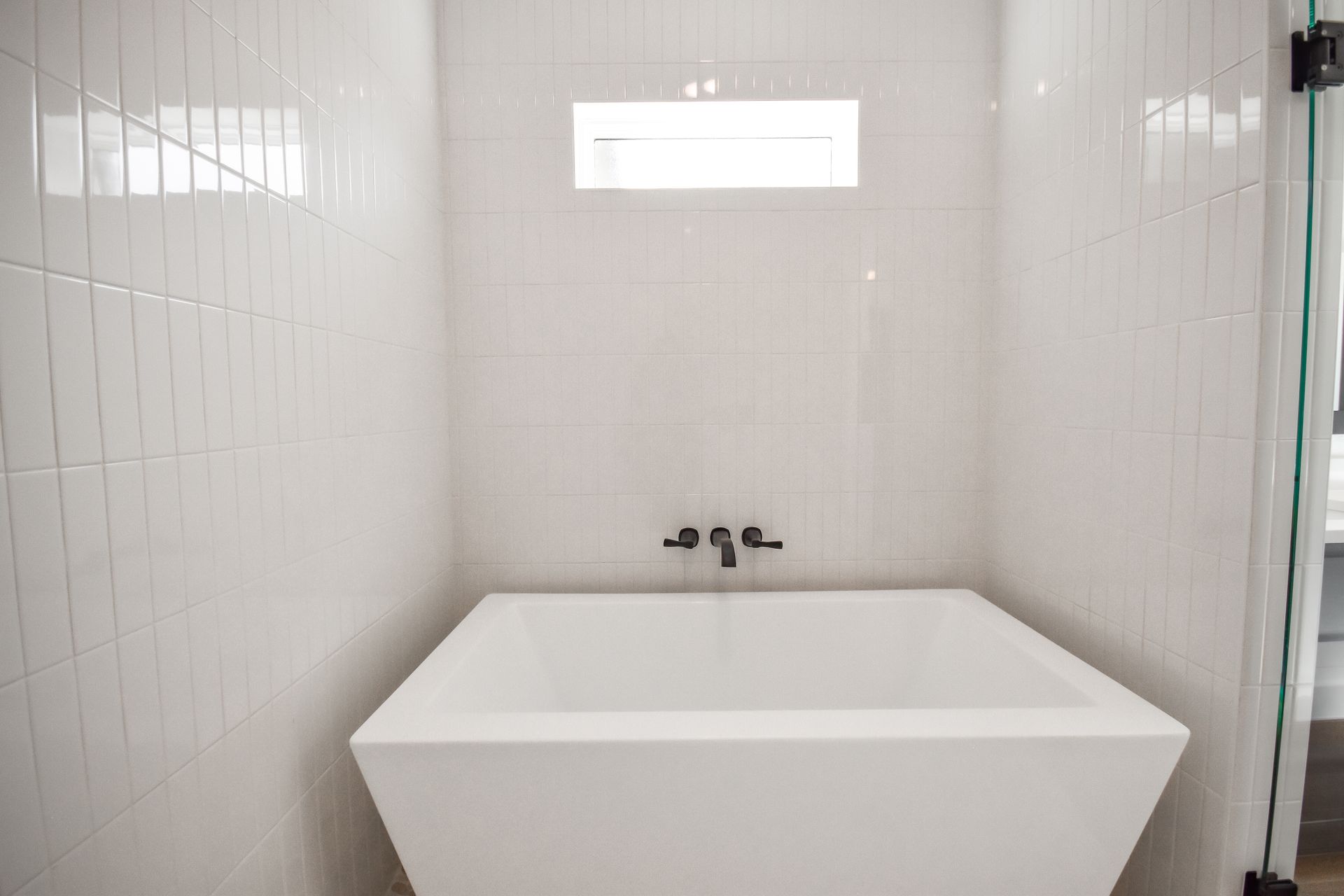 White modern bathroom with square tub, white tiled walls, and small window.