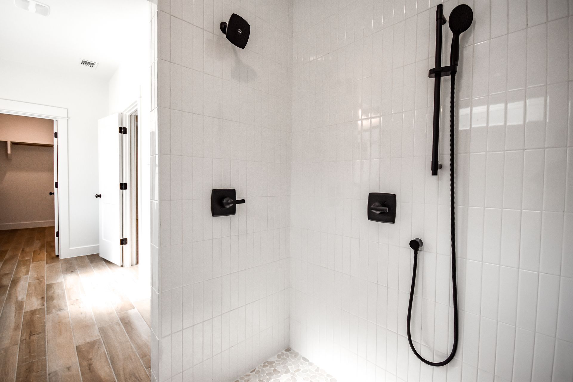White tiled shower with black fixtures and a hallway with a wooden floor.