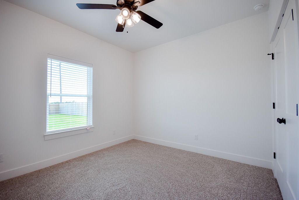 Empty room with tan carpet, white walls, window, ceiling fan, and closed white door.