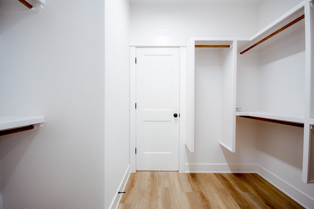Empty white closet with built-in shelving and door, wooden floor.