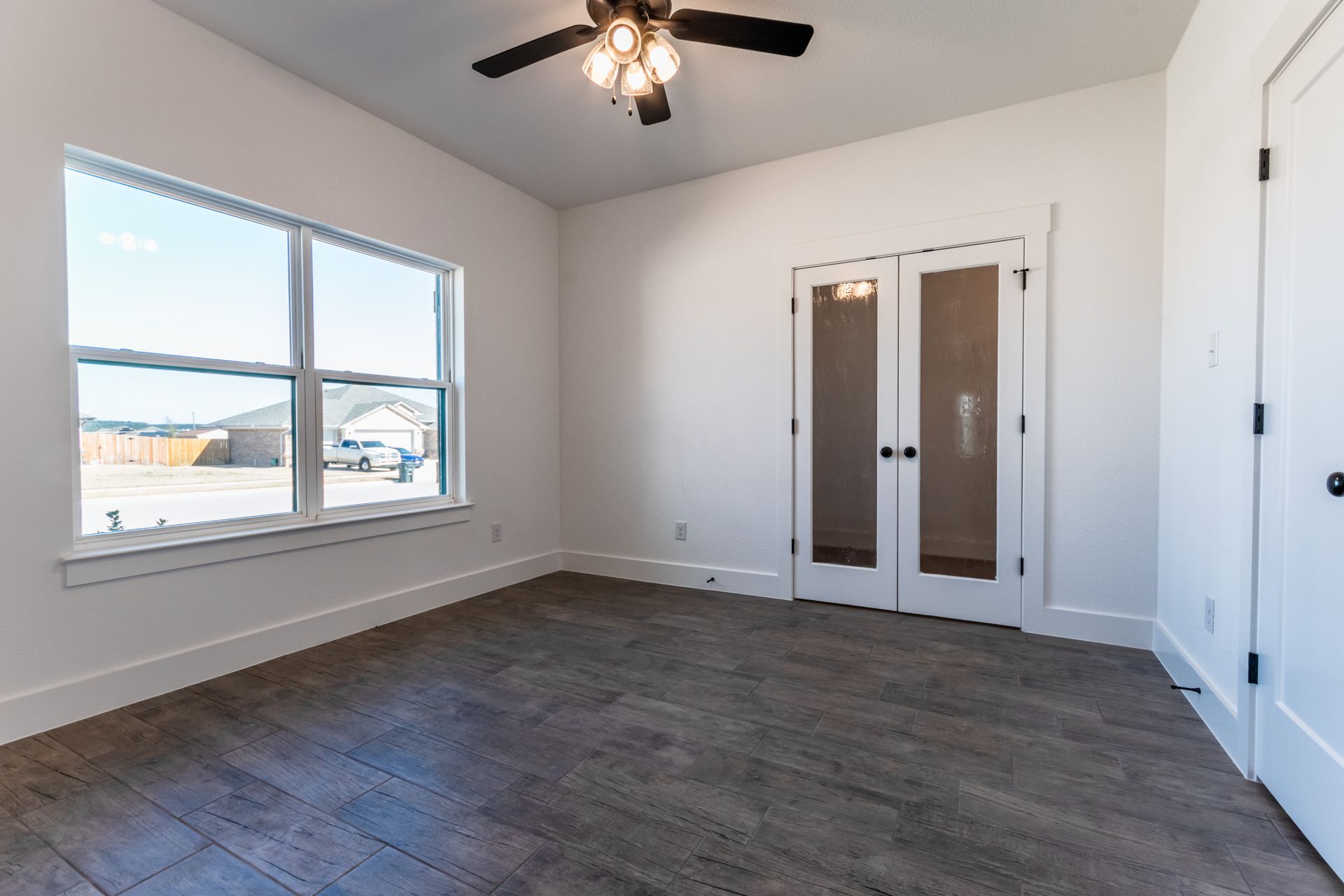 Empty room with large window, French doors, and ceiling fan. Gray flooring and white walls.