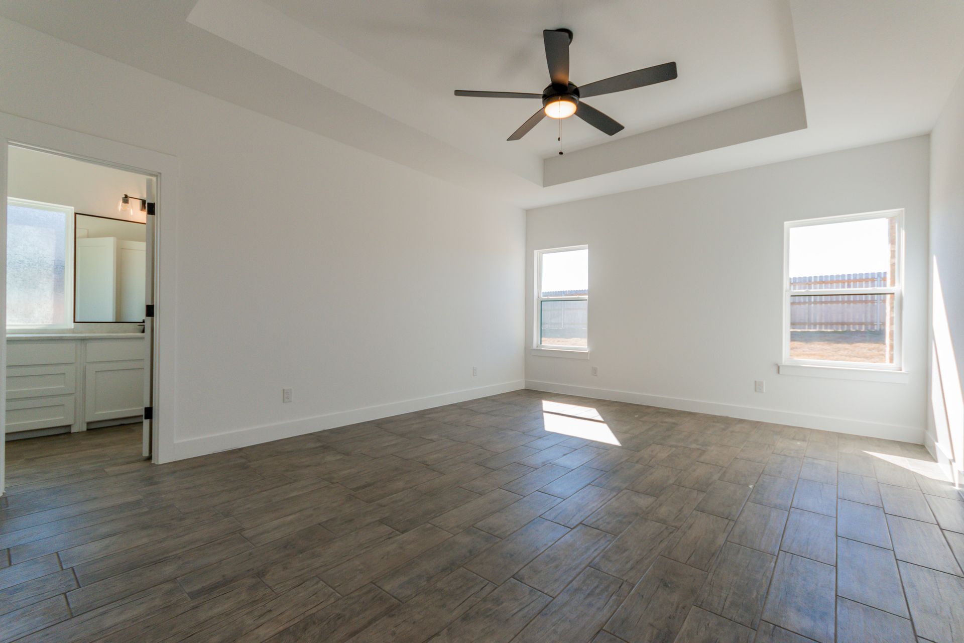 Empty bedroom with gray tile floor, white walls, and two windows. Bathroom doorway visible.