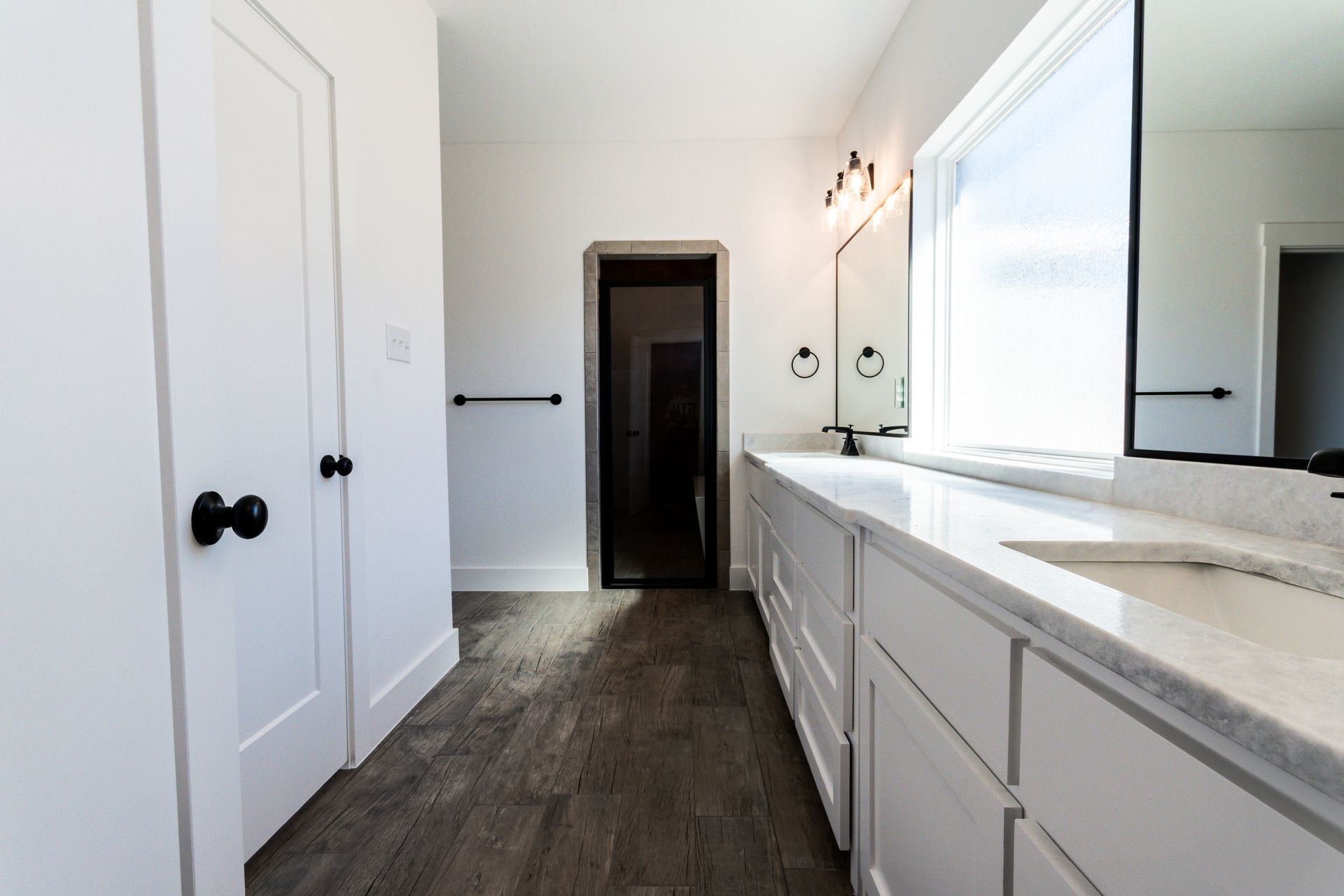 White bathroom with double vanity, dark floors, and a door to a dark room.